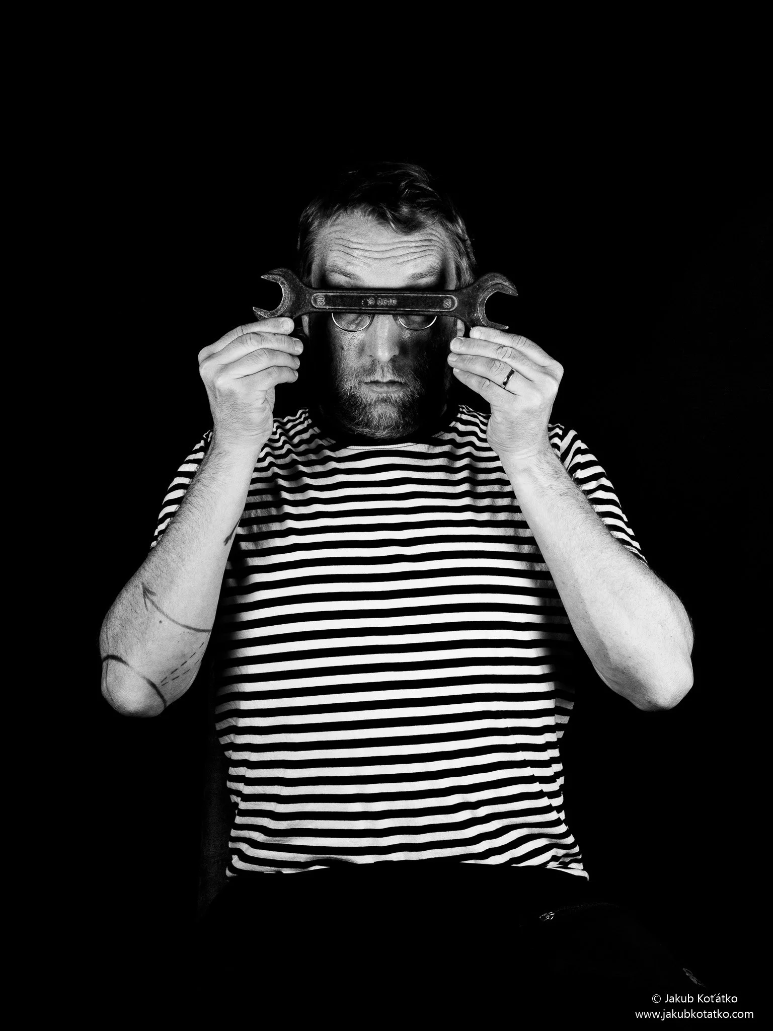 A man with glasses and a beard holding a wrench in front of his face, with a striped shirt, against a black background.