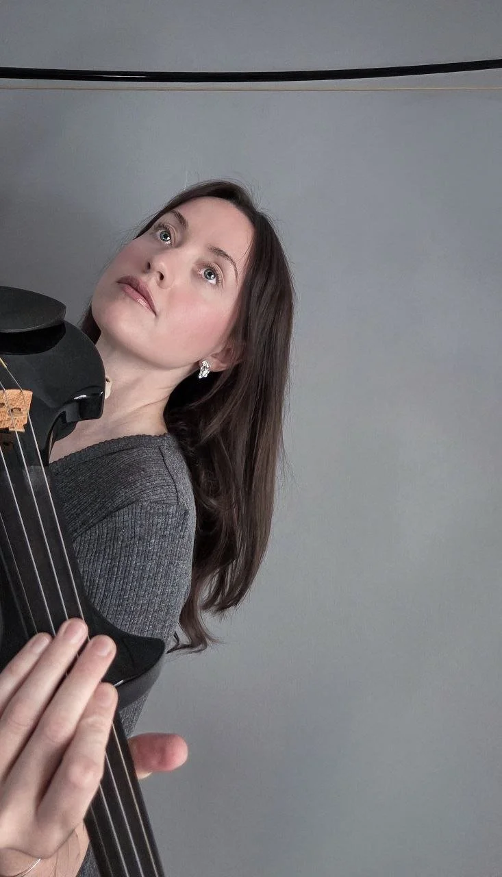 A woman with long brown hair and earrings holding a black violin, looking upwards, against a light gray background.