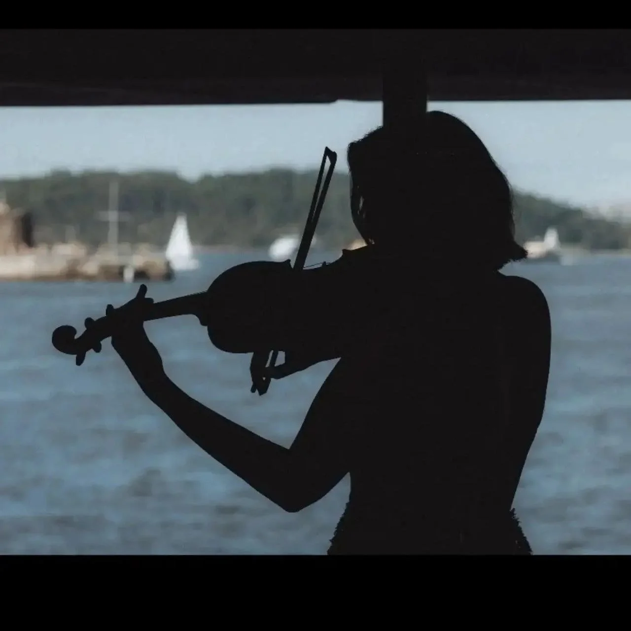 Silhouette of a woman playing the violin under a bridge with water and boats in the background.