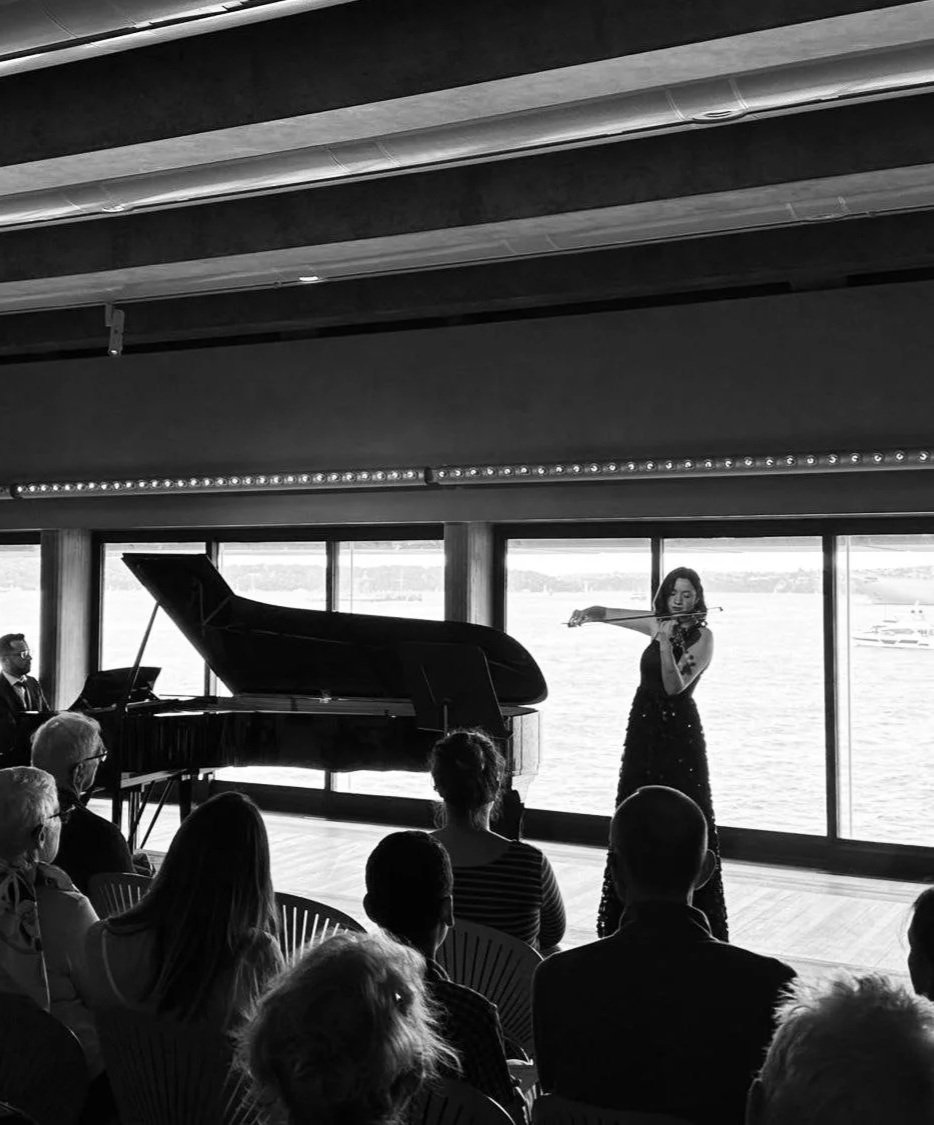 A woman playing the violin during a performance in a room with large windows overlooking a body of water, with an audience seated in front of her.