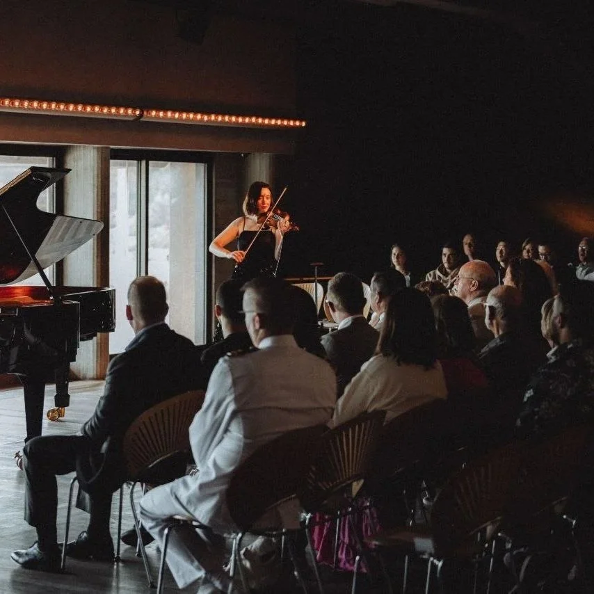 A woman playing the violin during a live music performance indoors, with an audience watching.