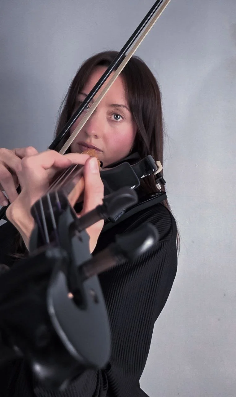 A woman playing a violin, looking directly at the camera, with the bow raised and waiting to play, against a plain gray background.