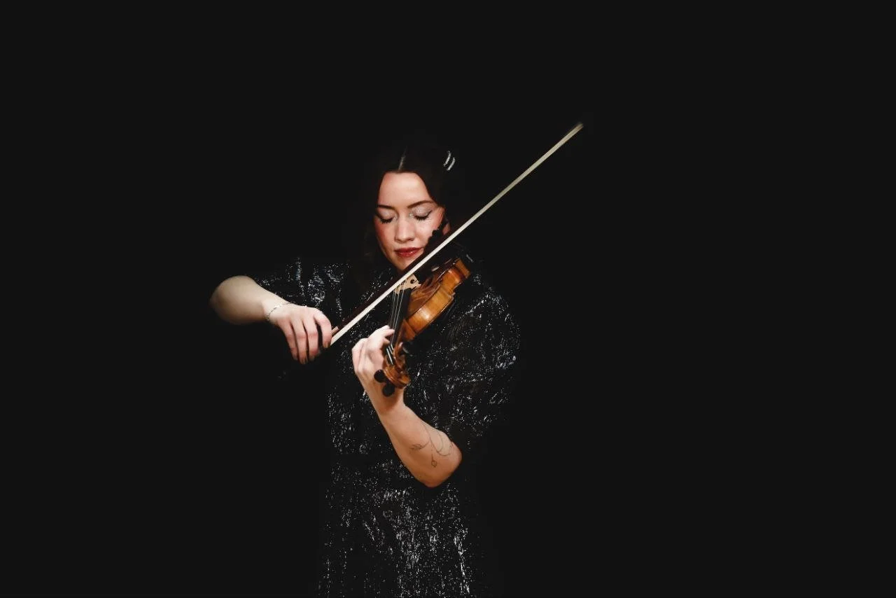 A woman with dark hair in a black, shiny, long-sleeved top playing a violin against a black background