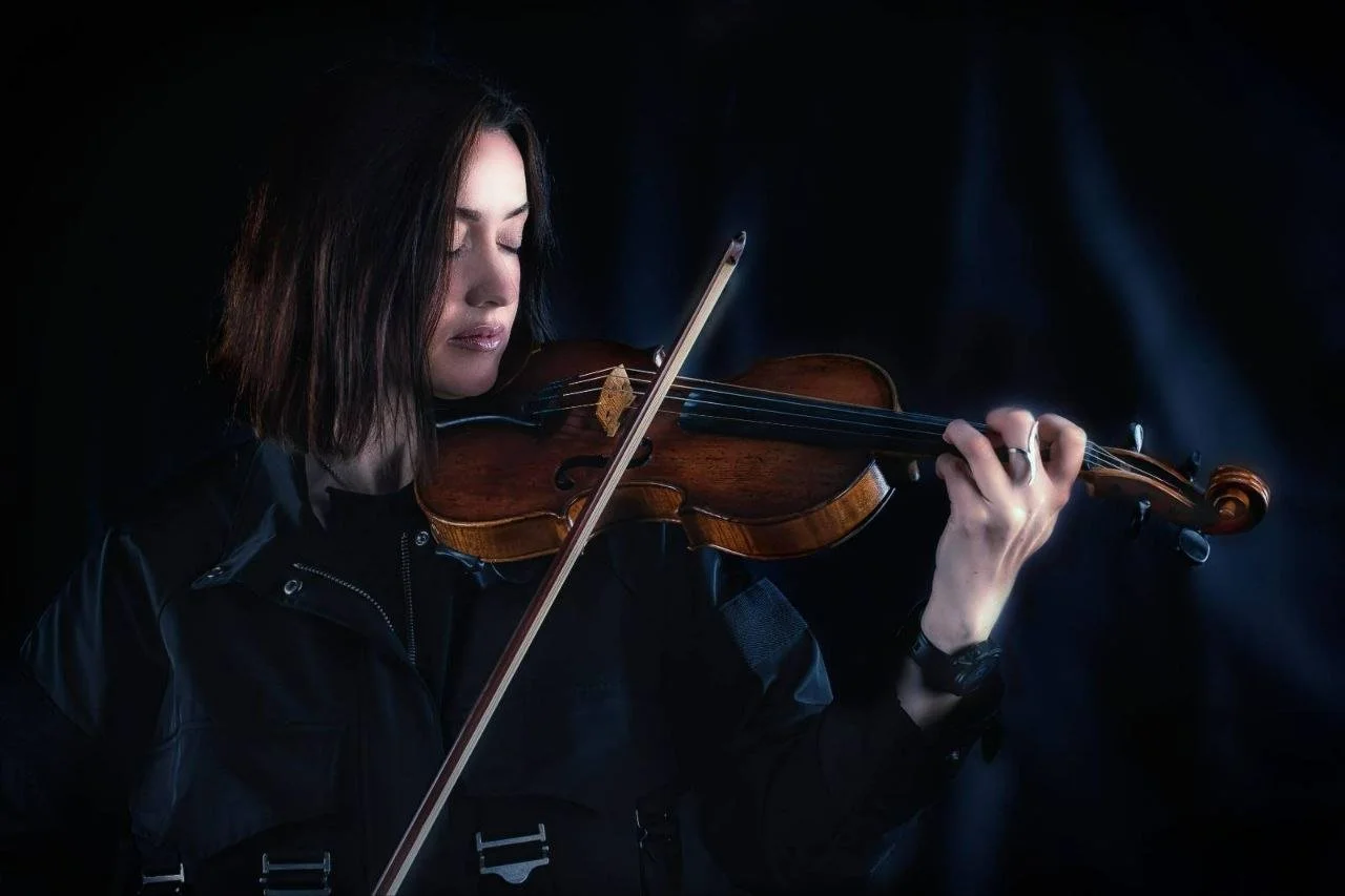 Woman with dark hair playing the violin in a dark setting with a black background.