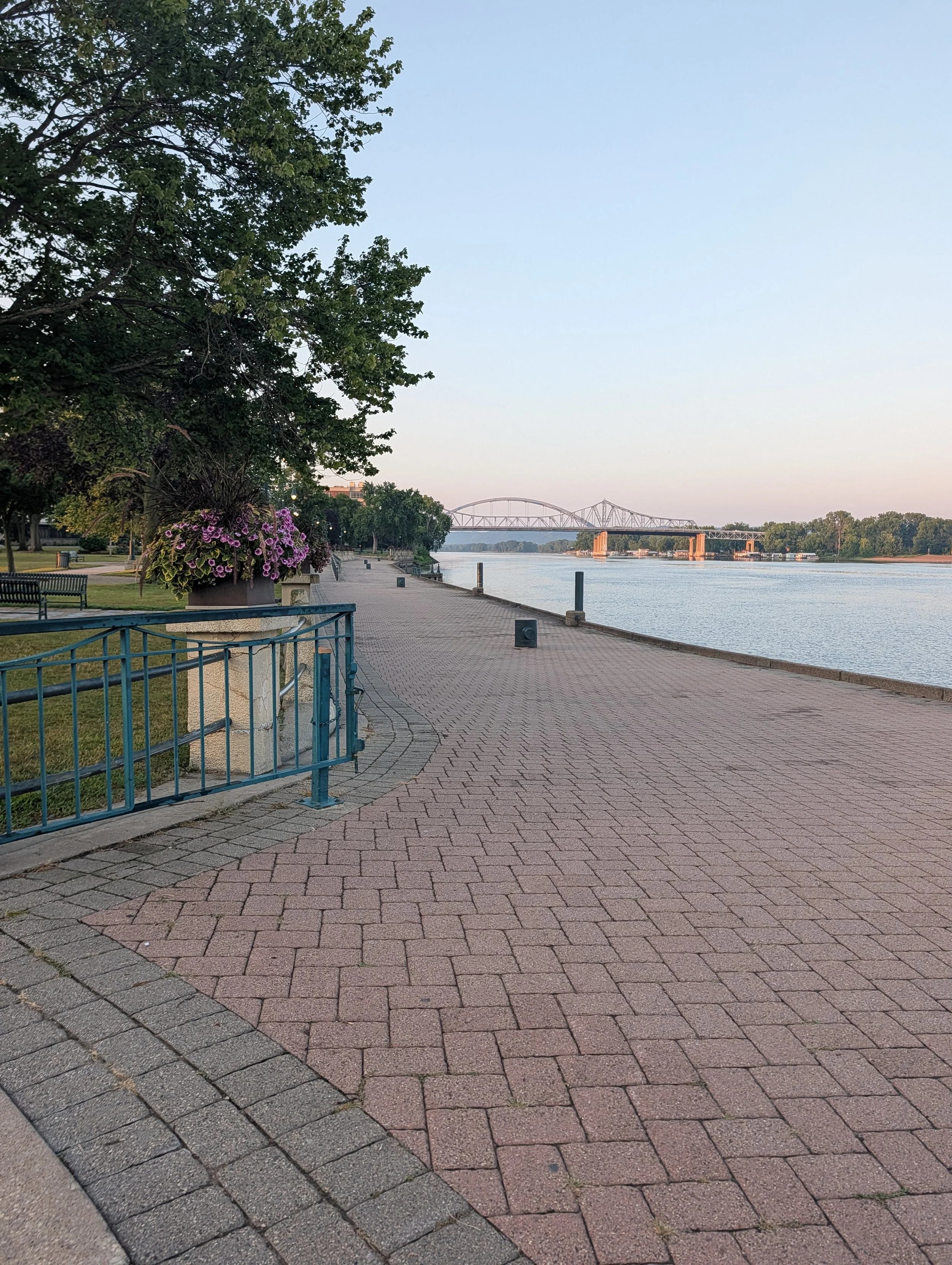 A riverside park walkway with brick pavers, a metal railing, benches, a large potted plant with pink flowers, and a view of a river and a bridge in the distance.