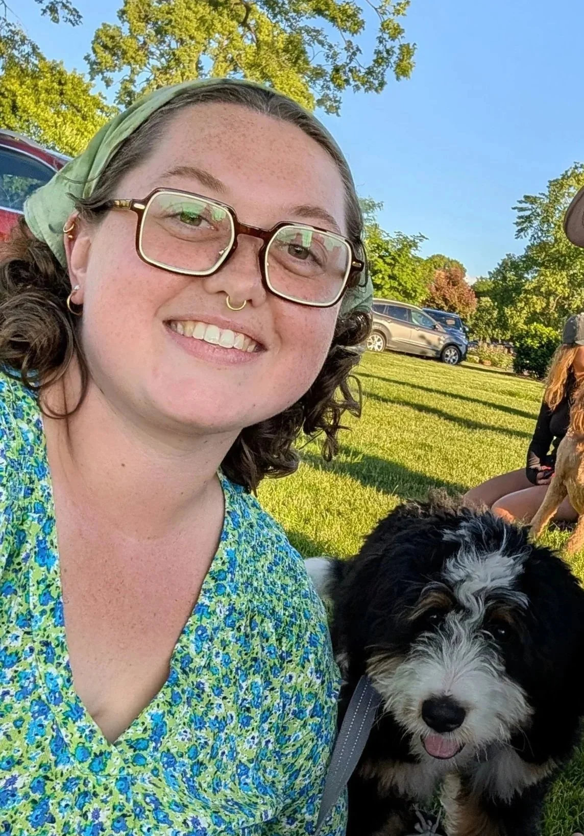 A woman with glasses and a green headscarf smiling in a park with a black and white puppy. Trees and parked cars are in the background on a sunny day.
