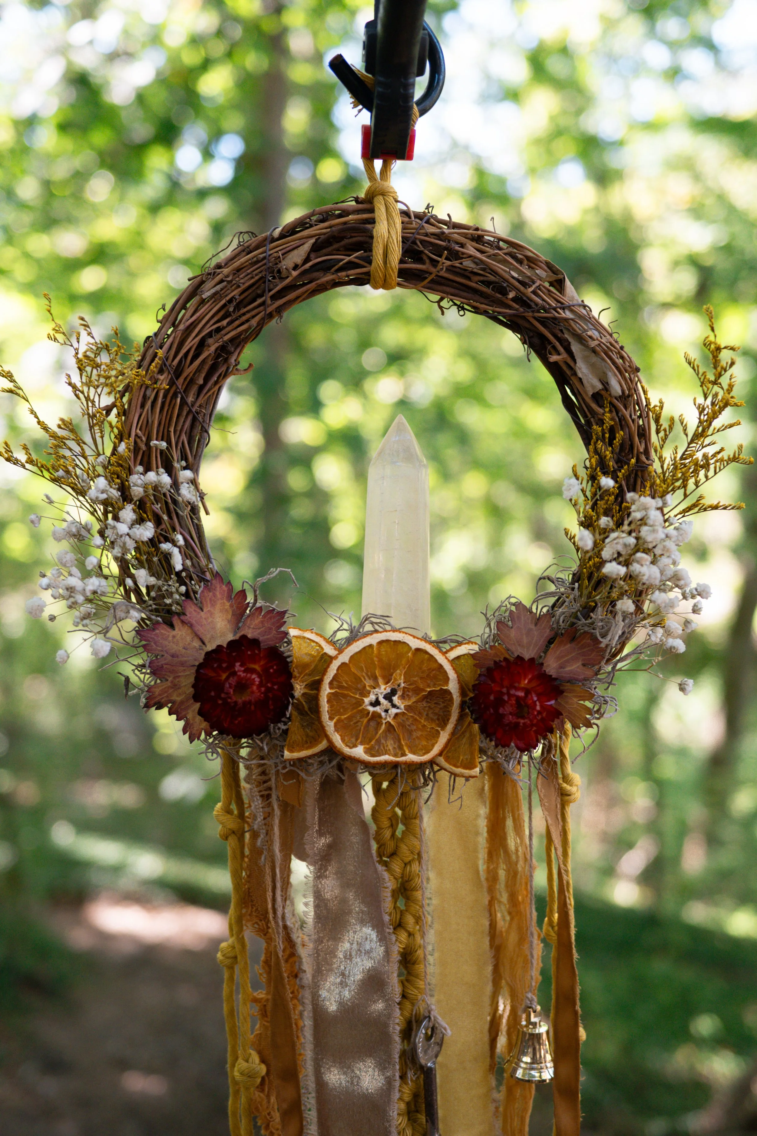 A decorative hanging wreath made of intertwined twigs adorned with dried orange slices, red flowers, white baby's breath, and yellow autumn leaves, with ribbons and a small bell hanging from the bottom, suspended outdoors against a blurred background of green trees and sunlight.