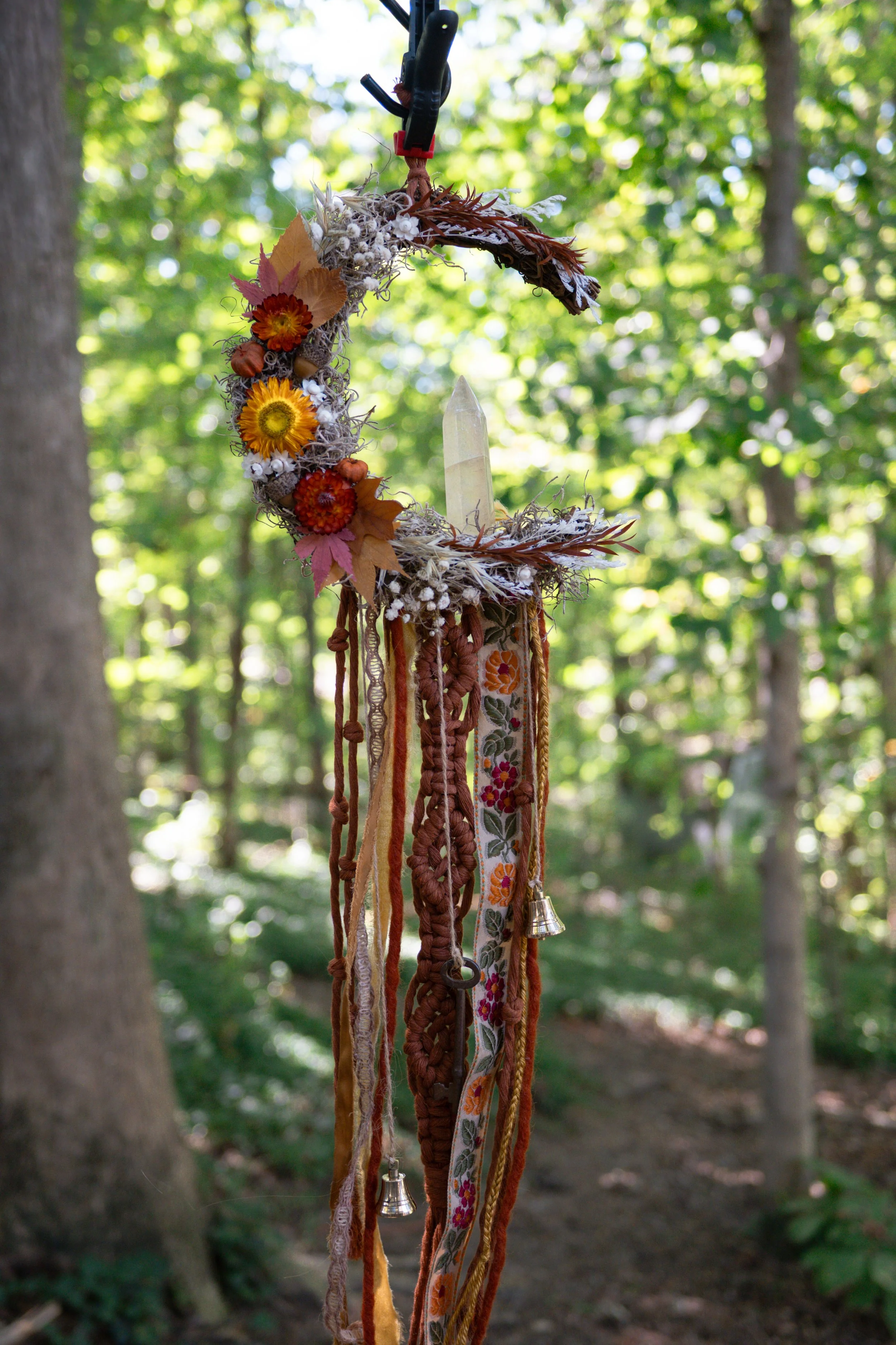 A decorative hanging ornament with dried flowers, leaves, and ribbons, featuring a central clear crystal points, hanging in a wooded outdoor setting with green trees in the background.