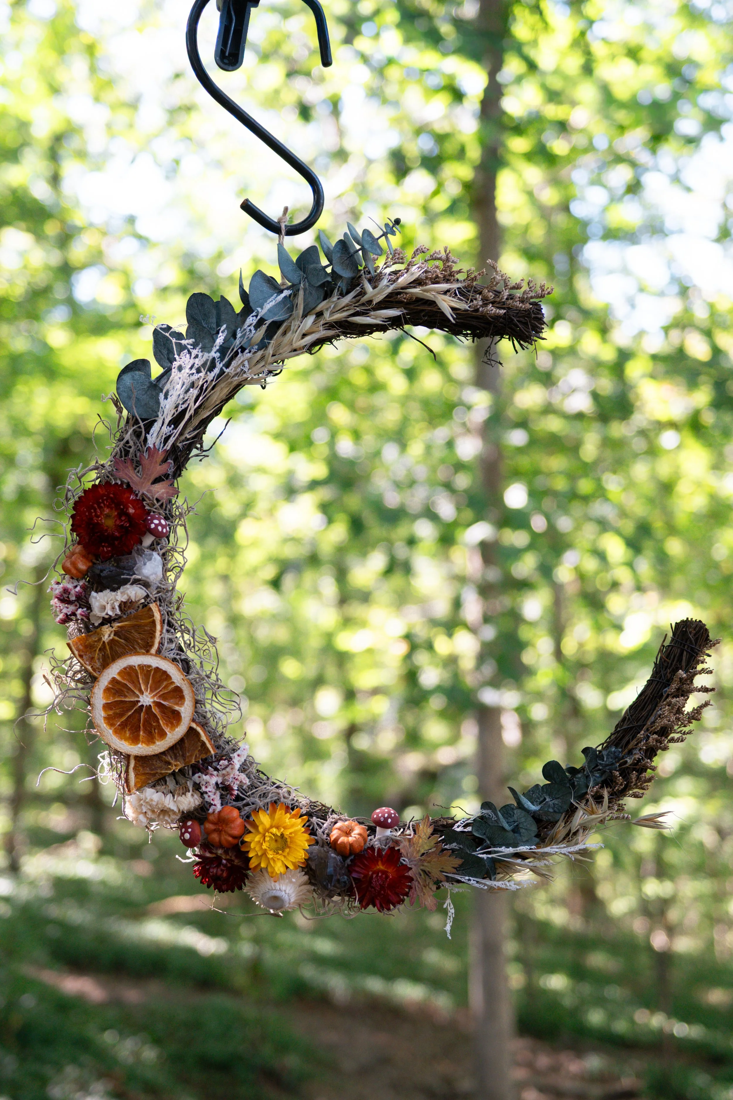 Close-up of a crescent-shaped floral arrangement with dried orange slices, yellow and red flowers, mushrooms, and various dried plant materials, hanging outdoors with green trees in the background.