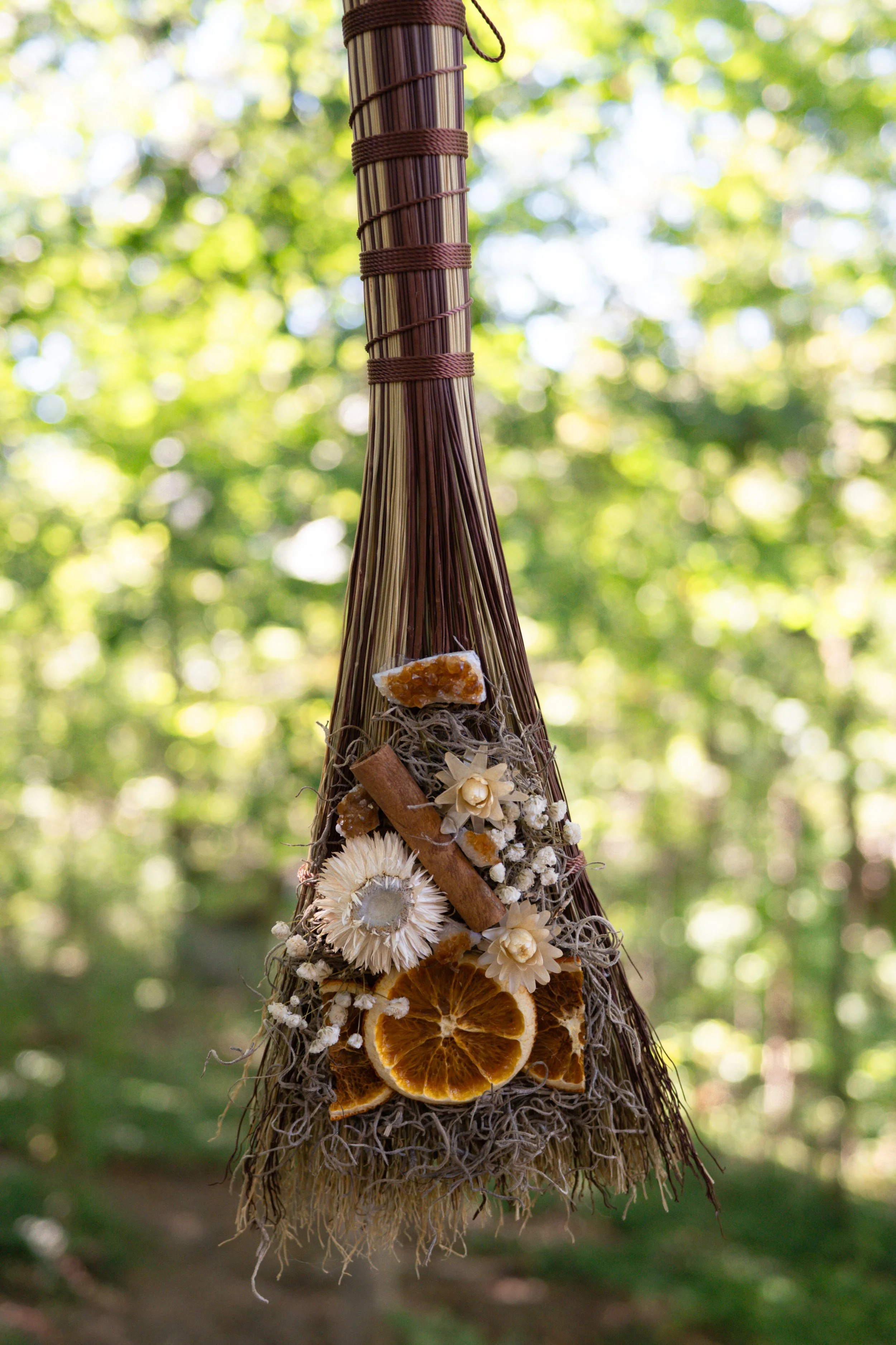 Decorative hanging ornament made of dried citrus slices, flowers, and natural materials, hanging outdoors with a blurred background of green trees and sunlight.