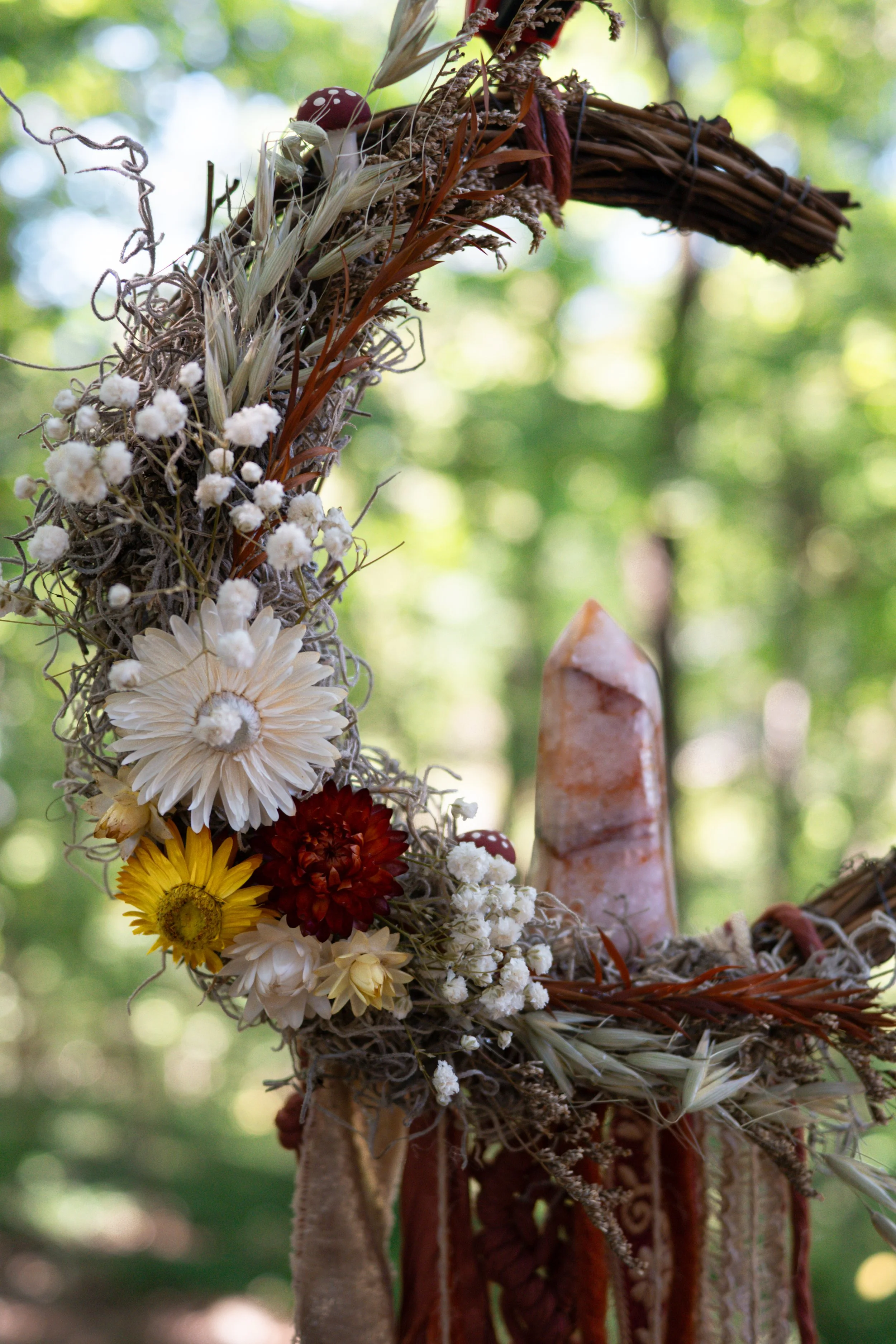 A close-up of a handmade wreath decorated with dried flowers, grasses, and small mushrooms, hanging outdoors against a blurry background of green trees and light.