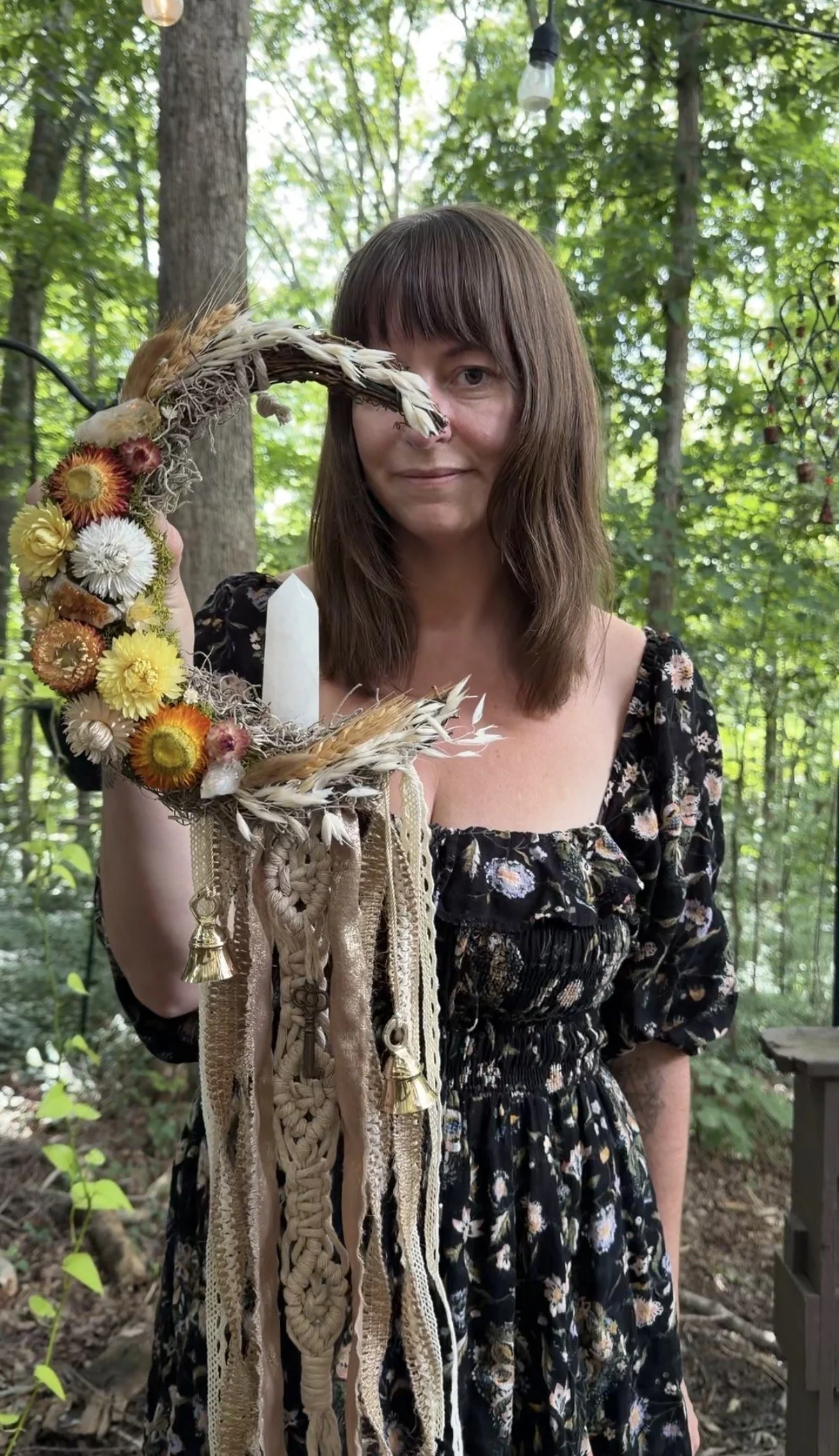 A woman with brown hair in a black floral dress holding a decorative crescent-shaped wreath made of dried flowers, feathers, and hanging ribbons in a wooded outdoor setting.