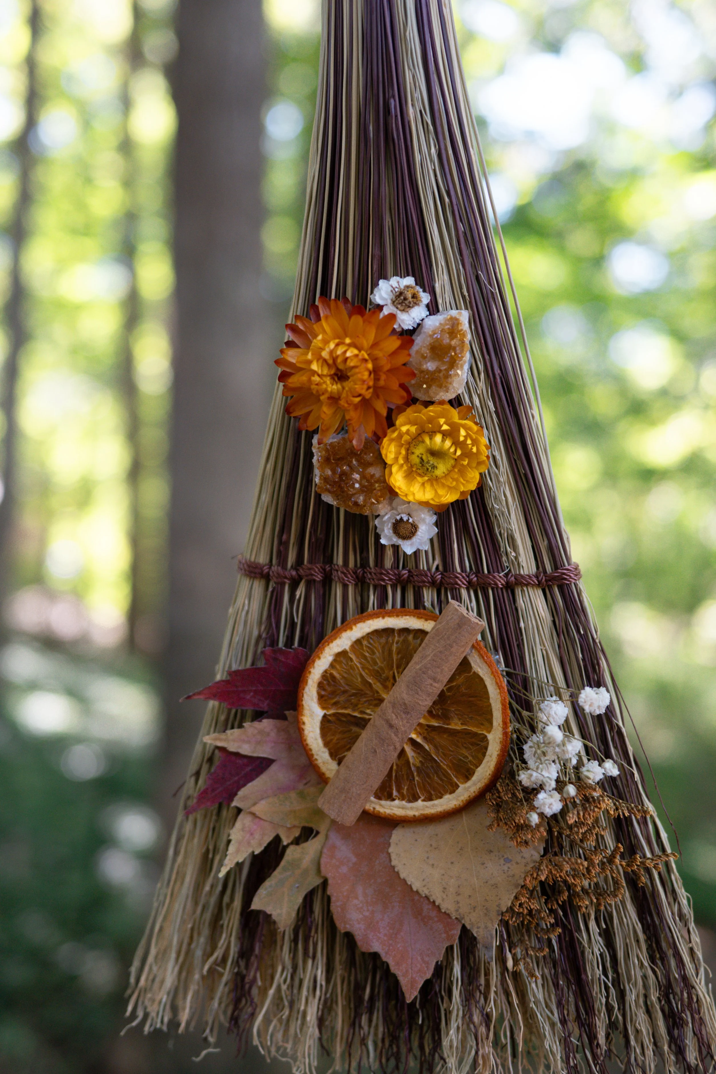 A decorative arrangement on a woven straw object featuring dried orange slices, cinnamon sticks, and various dried flowers and leaves, set outdoors with a blurred green forest background.