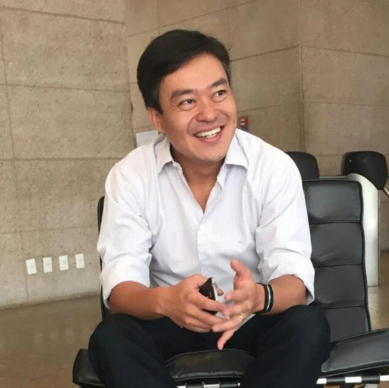 Marcio Kawashima sentado em uma cadeira de couro preto, usando camisa branca de mangas longas, dentro de um ambiente com paredes de pedra e tomadas elétricas visíveis na parede ao fundo.