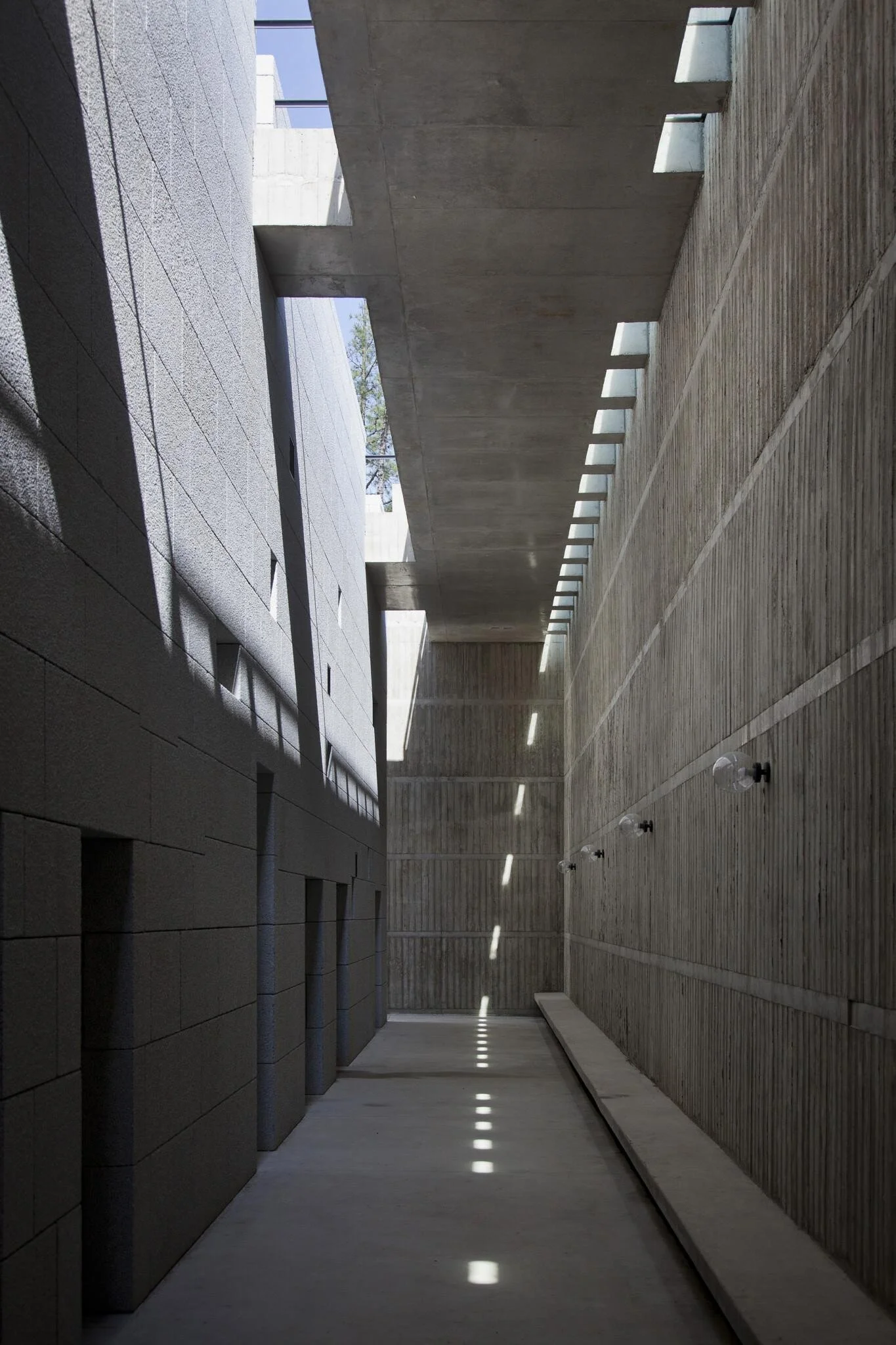 Corredor interno de edifício moderno com paredes de lajes de concreto, piso de mármore e iluminação natural entrando por aberturas no teto.