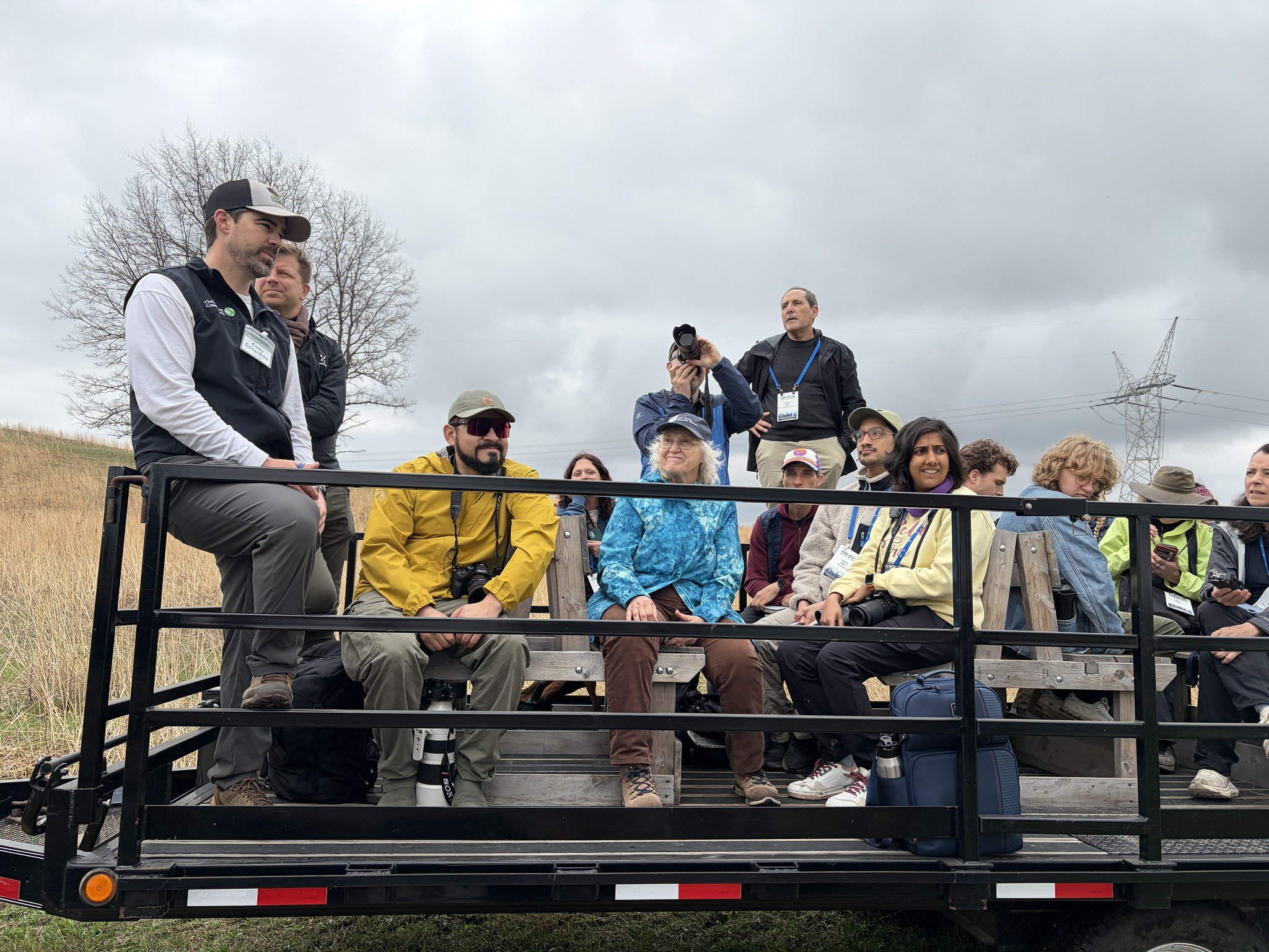 Group of people sitting and standing on an outdoor flatbed trailer, some with cameras, during cloudy weather, with leafless trees and power lines in the background.