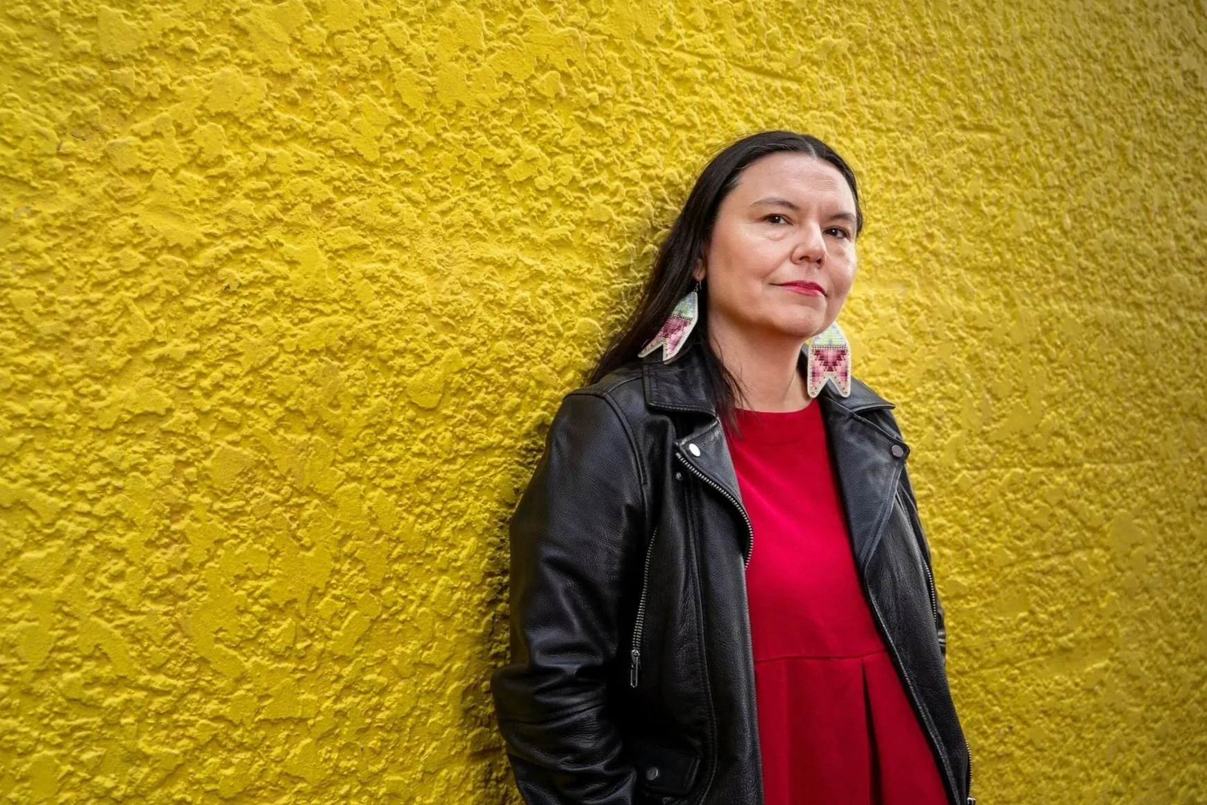 A woman standing against a bright yellow textured wall, dressed in a red top and black leather jacket, wearing colorful beaded earrings.