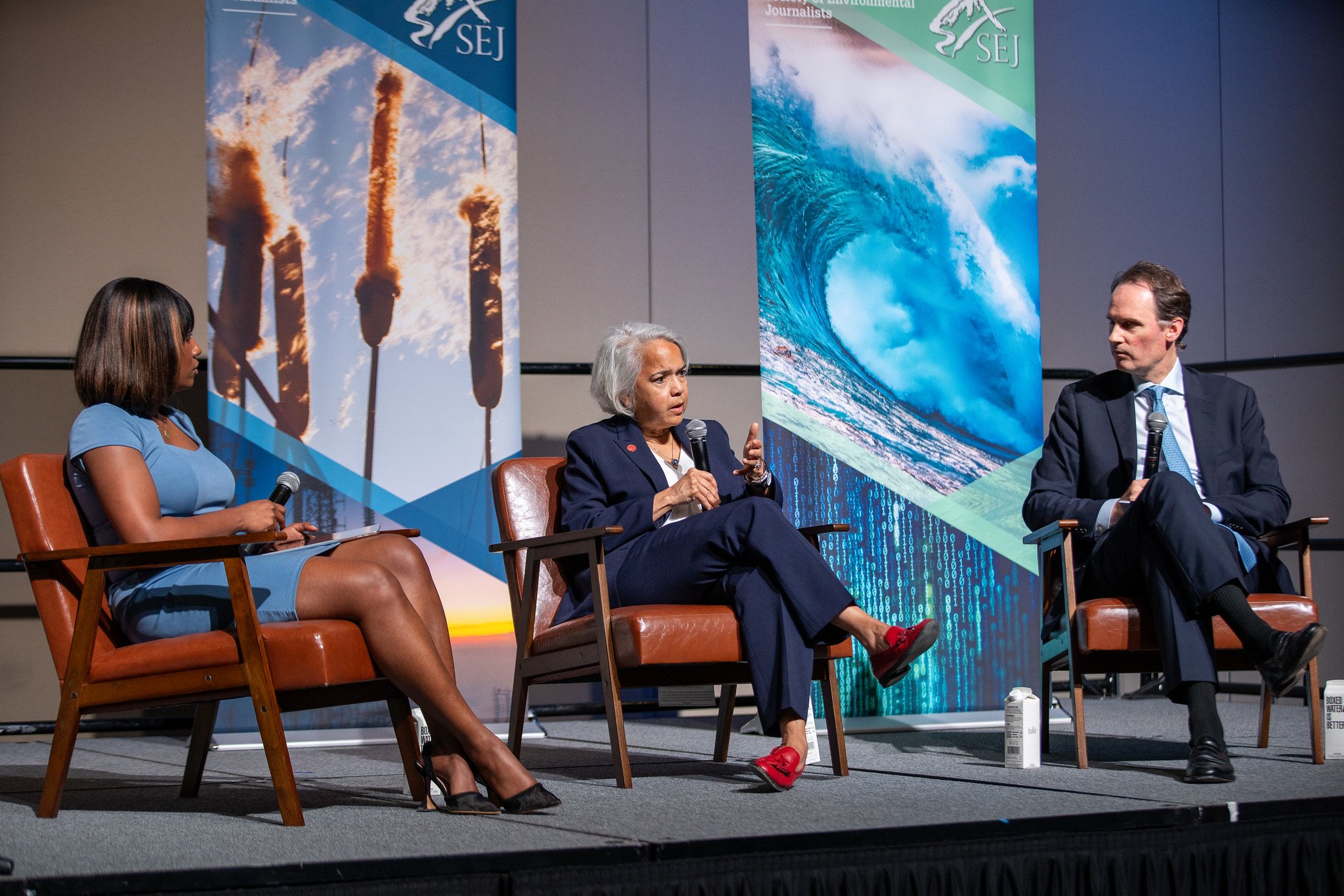 A panel discussion featuring three individuals seated on stage with large banners behind them. The banners display images of pollution and waves and have the SEJ logo. The woman in the middle is speaking into a microphone, wearing a dark blazer and red shoes. The woman on the left is wearing a blue dress, and the man on the right is dressed in a suit with a light blue tie, holding a microphone.