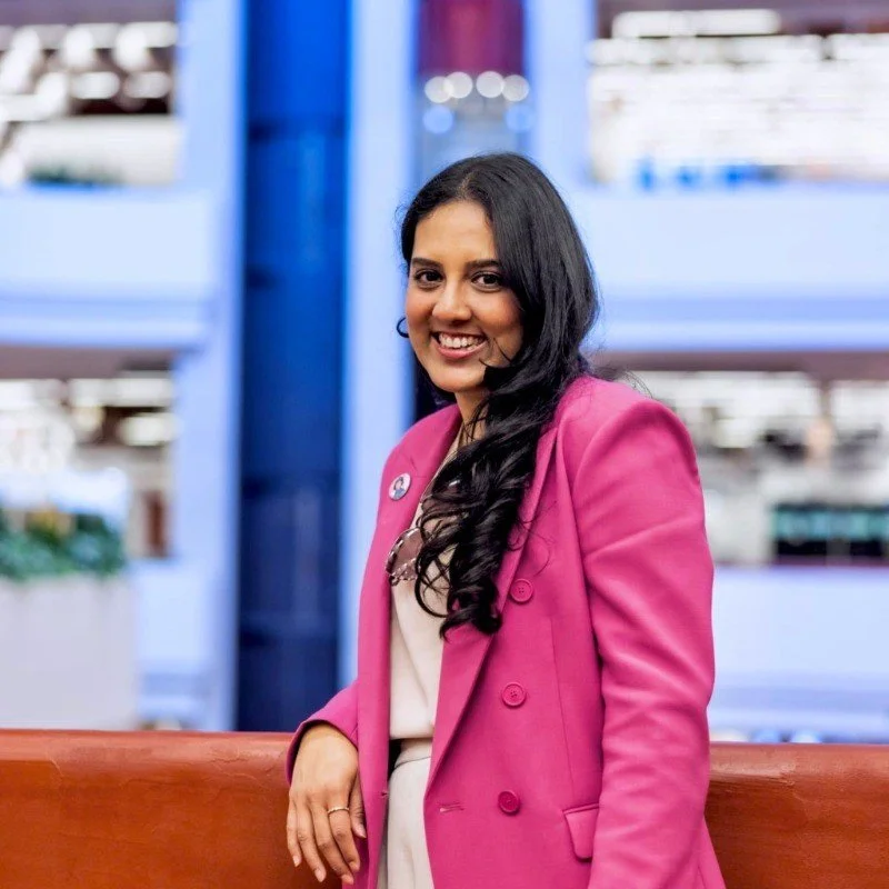 A woman with long dark hair smiling, wearing a pink blazer over a white top, in a modern indoor setting.