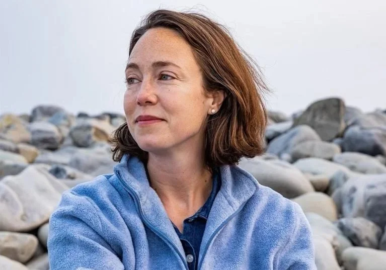 A woman with shoulder-length brown hair wearing a blue fleece jacket, standing outdoors against a background of rocks.