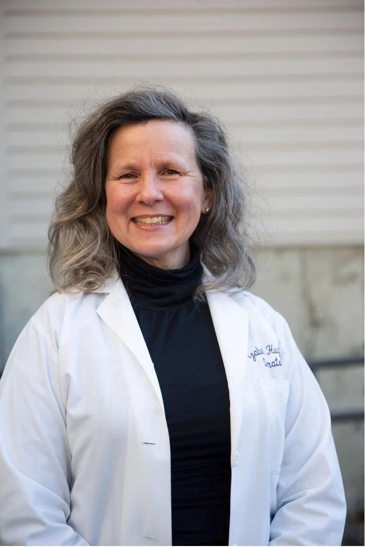 A smiling woman with shoulder-length wavy gray hair, wearing a white lab coat over a black turtleneck, standing outdoors in front of a light-colored building with horizontal siding.