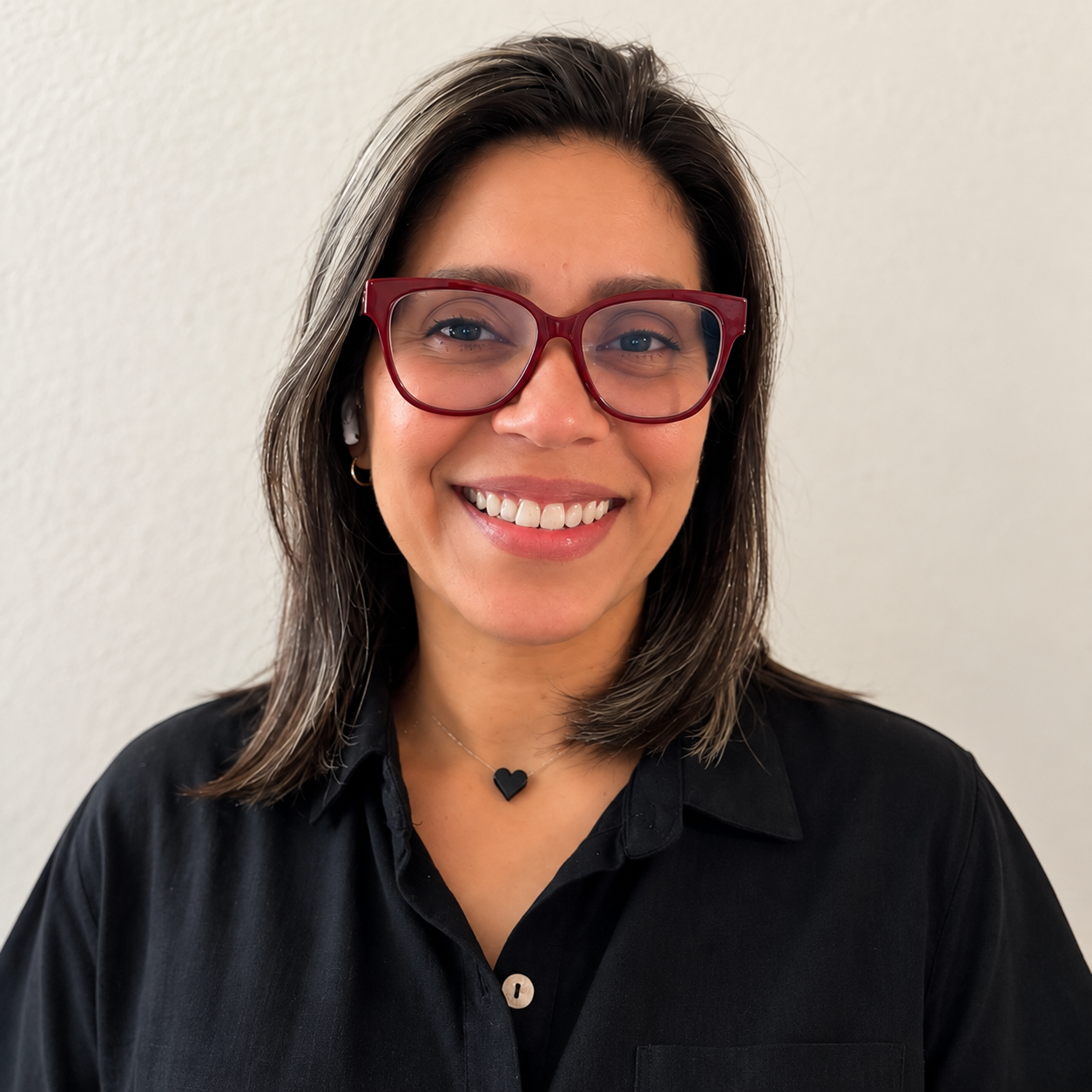 A woman smiling, wearing red glasses, a black shirt with a collar, a small black heart necklace, and AirPods, standing against a light-colored wall.