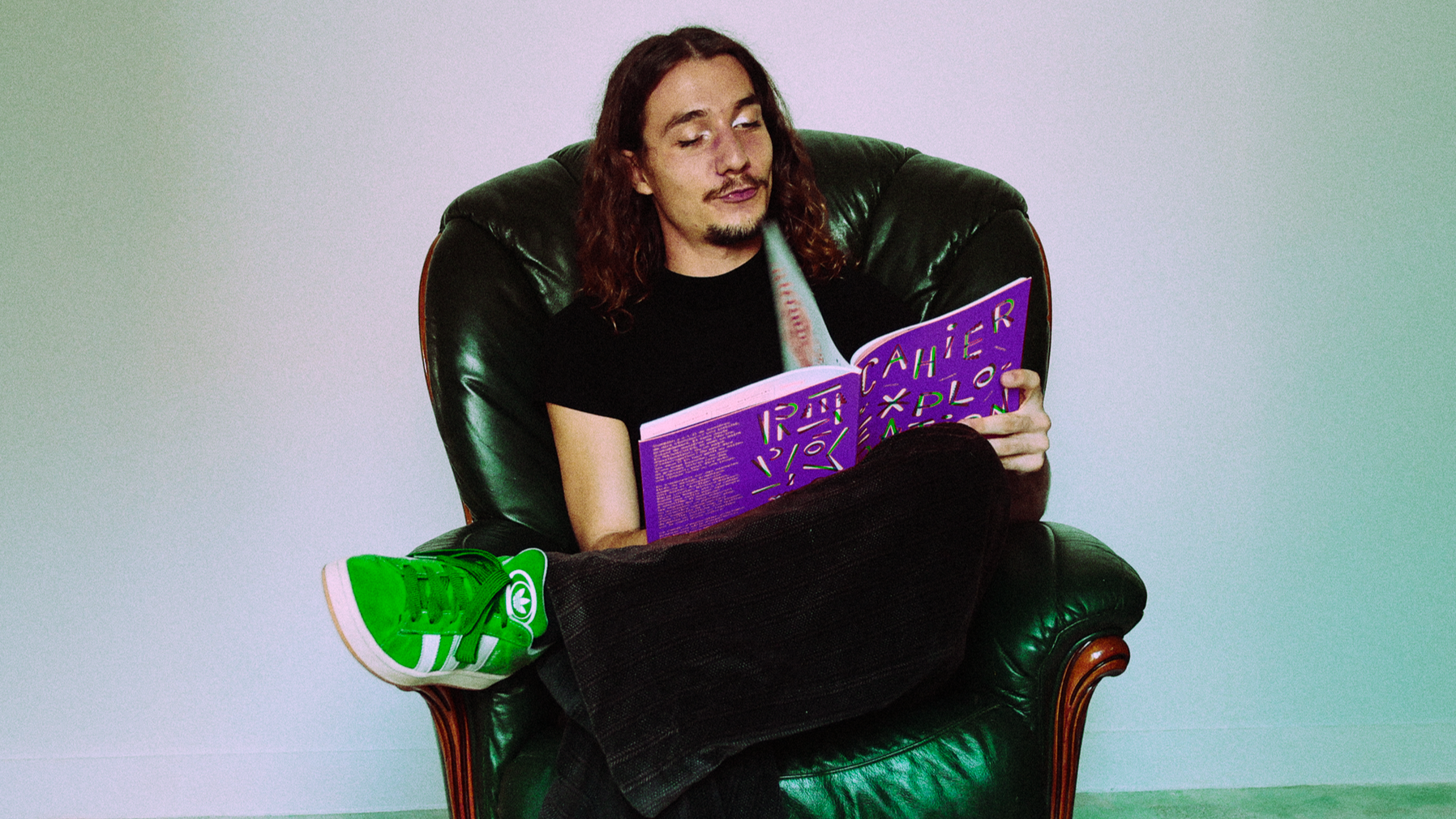 Un homme assis dans un fauteuil en cuir vert foncé, rieur, lisant un magazine violet avec des motifs colorés.