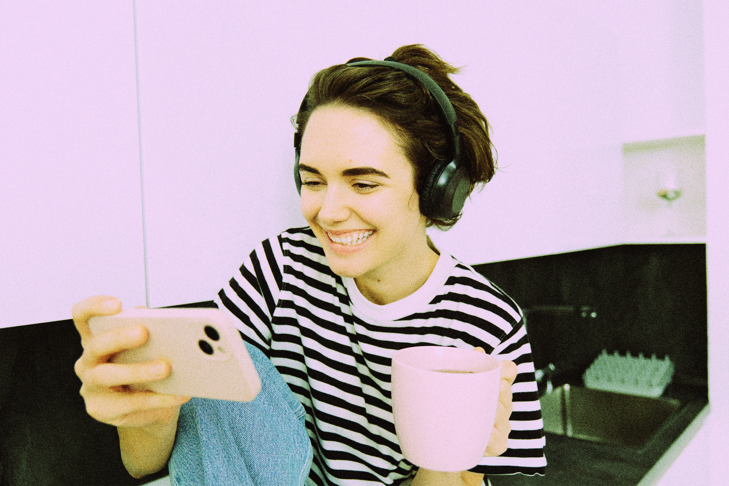 Une jeune femme souriante portant un casque écoute de la musique, regardant son téléphone, avec une tasse rose sur la table devant elle, dans un intérieur sombre.