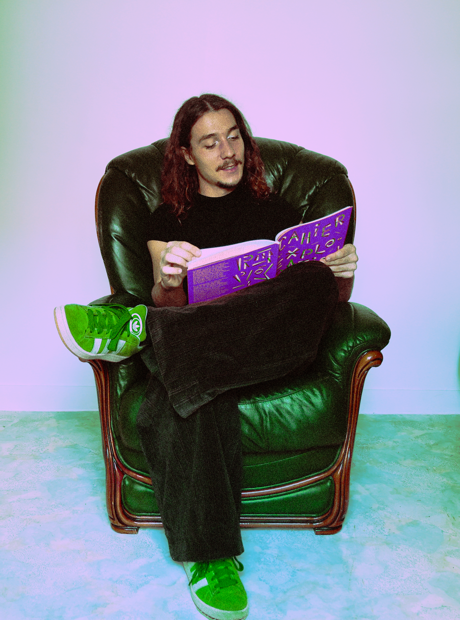 Une personne à cheveux longs et bouclés, assise dans un fauteuil en cuir vert foncé, lit un magazine violet avec des textes et des dessins. Elle porte un t-shirt noir, un pantalon noir, une chaussure verte avec des lacets blancs, et une autre chaussure visible à côté d’elle. Le fond est une surface blanche.