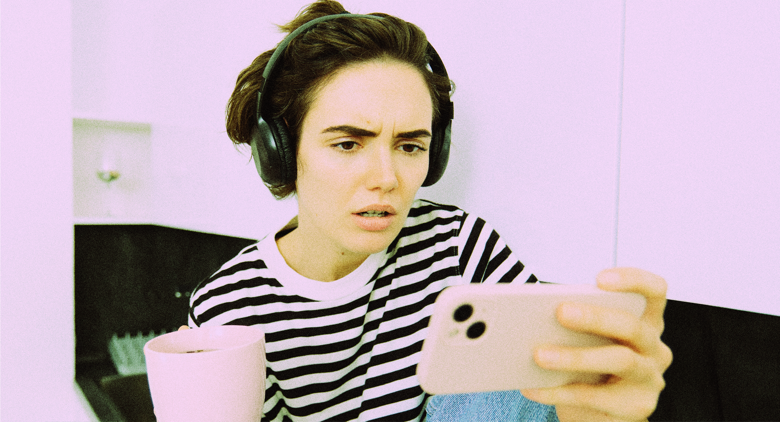 Une jeune femme avec des cheveux courts, portant un casque d'écoute et une chemise à rayures noires et blanches, regarde son téléphone portable avec une expression de surprise ou de confusion. Elle est assise à un bureau, avec une tasse rose sur la table.