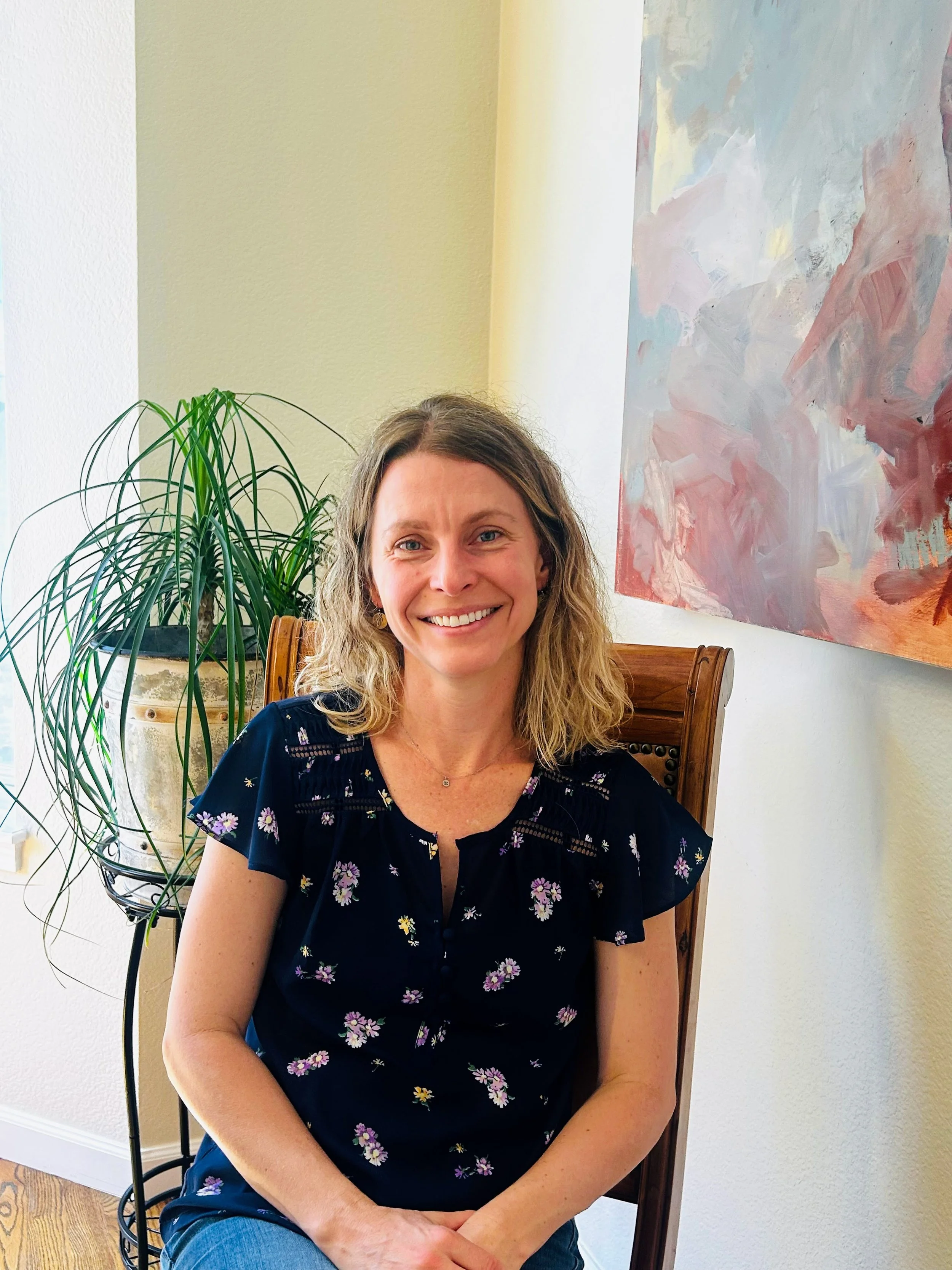A woman with curly blonde hair and blue eyes sitting on a wooden chair, smiling, wearing a navy blue dress with floral pattern, in a room with light-colored walls, a green plant, and an abstract painting.