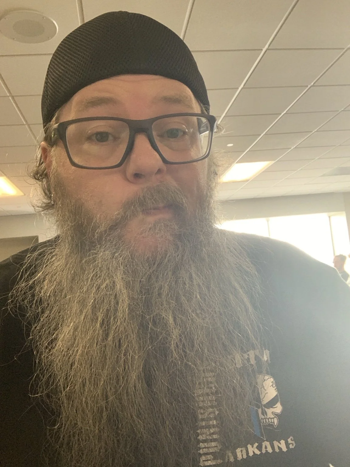 A man with glasses, a black beanie, and a long gray beard taking a selfie in an indoor setting with a ceiling with lights and other people in the background.