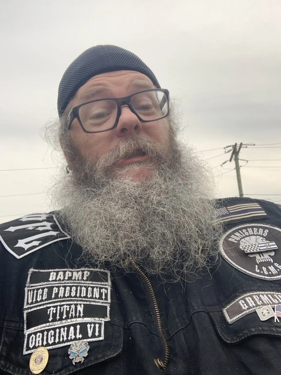Close-up of a man with glasses, a beanie hat, and a long gray beard wearing a black vest with patches.