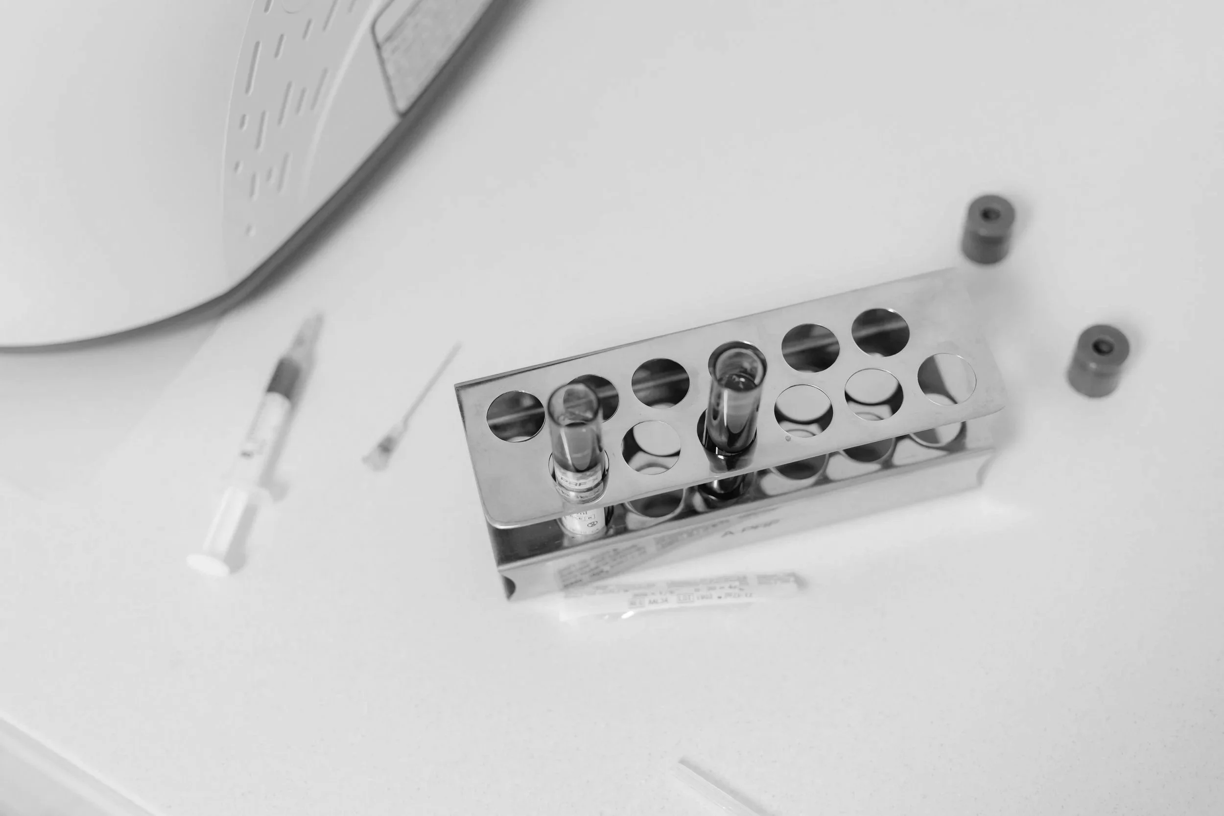 Close-up of a laboratory test tube rack with glass test tubes inside, set on a white lab counter. There are a few small laboratory tools or items nearby.