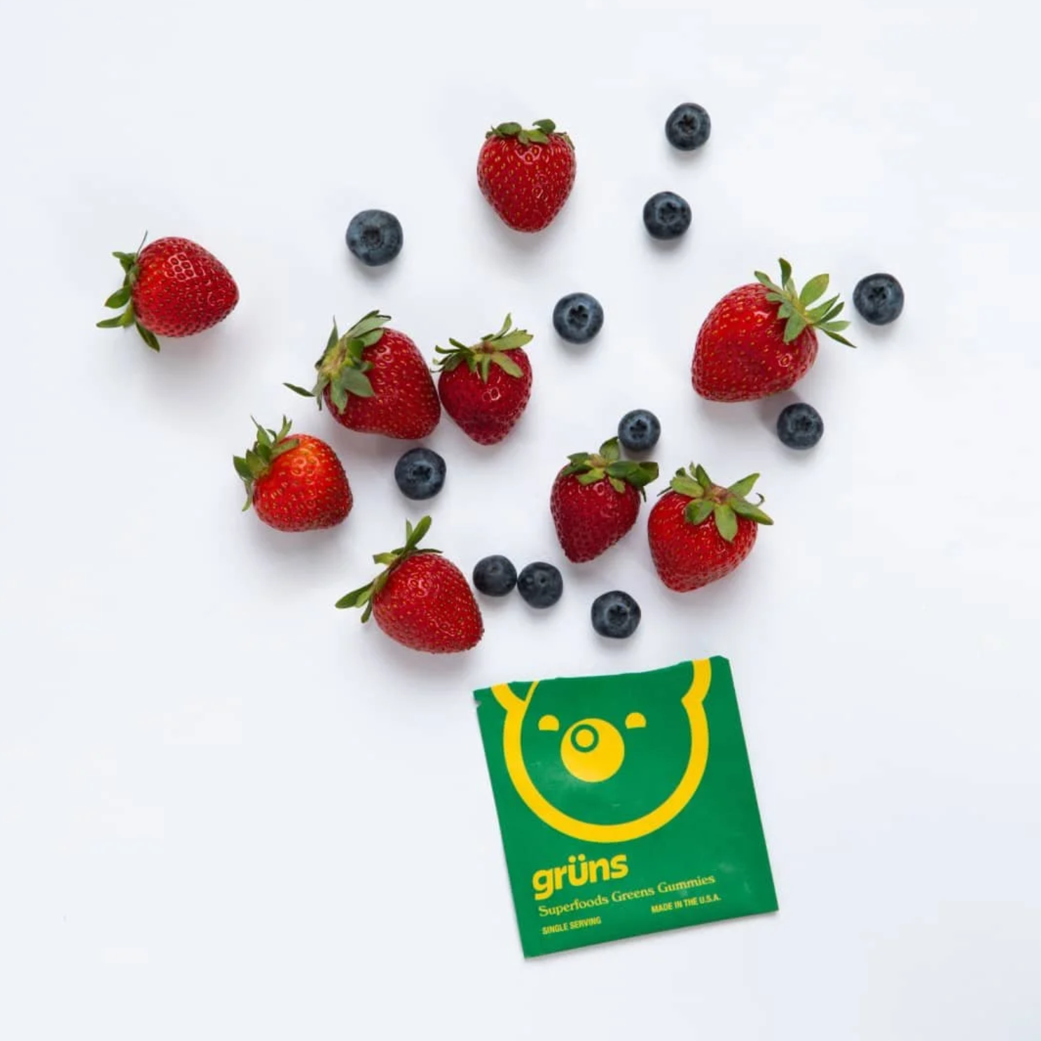 Fresh strawberries and blueberries scattered on white background with a green packet labeled 'grüns' superfoods gummies.