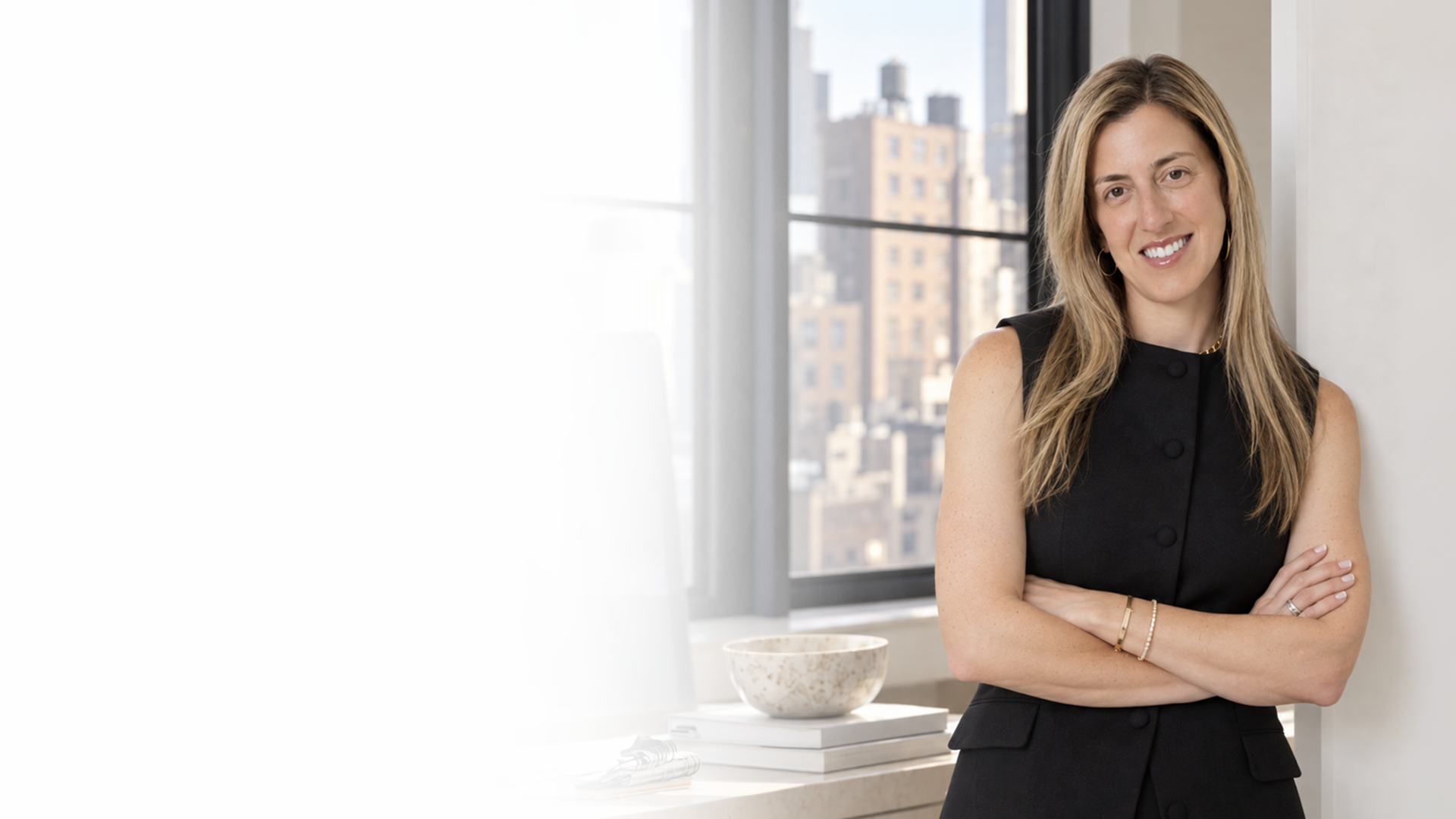 A woman with long blonde hair standing in an office with large windows showing a city skyline, wearing a black sleeveless top, smiling with her arms crossed.
