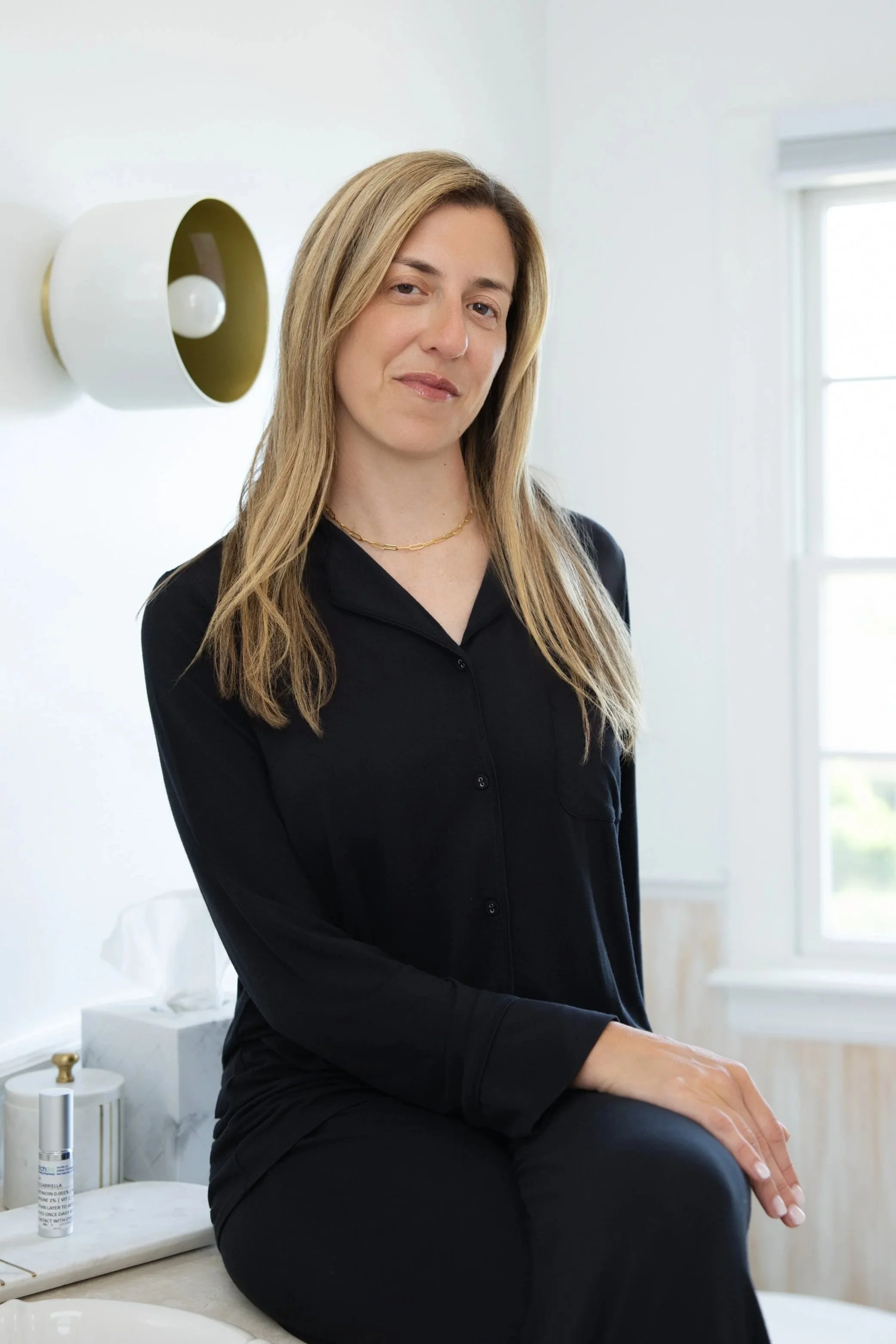 Woman with blonde hair wearing a black shirt sitting in a bright room with a window behind her, a modern white wall-mounted light fixture, and skincare products on a marble surface.