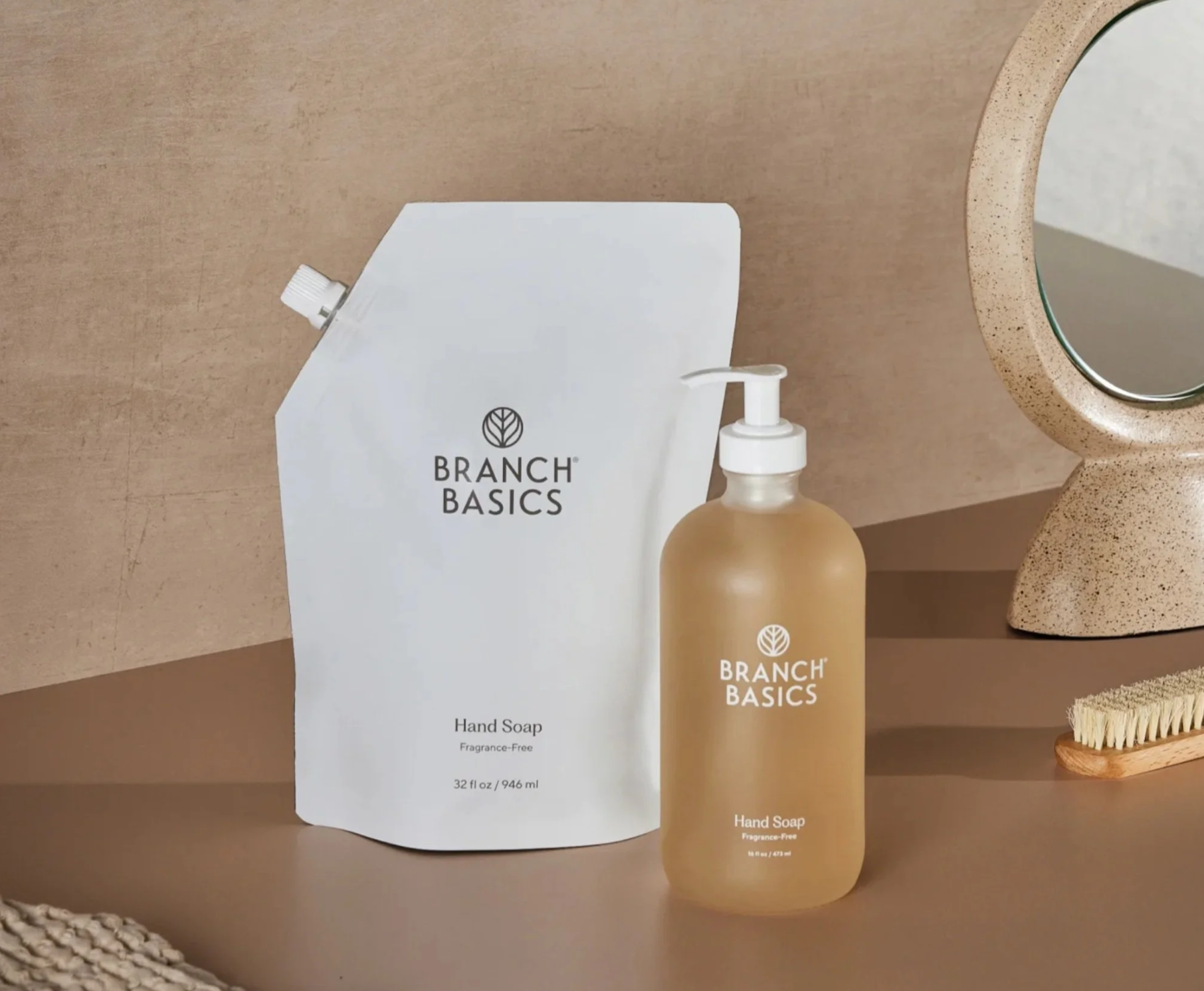 A beige countertop with a round mirror, a bottle of Branch Basics hand soap, a refill pouch of Branch Basics hand soap, a scrub brush, and a small cleaning brush.