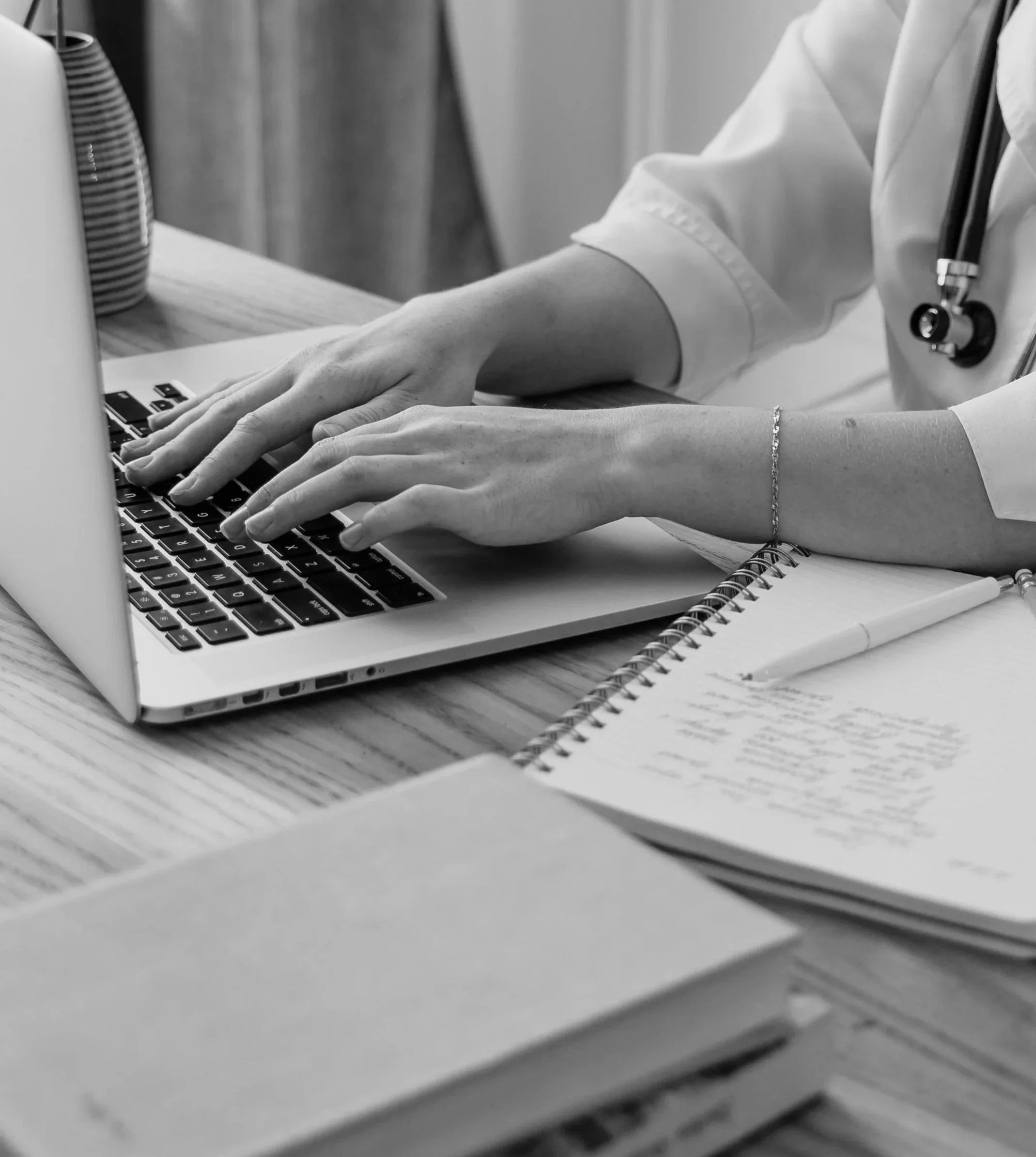 Close-up of a person's hands typing on a laptop keyboard at a desk with notebooks and a notebook to the side, wearing a white shirt and a stethoscope around their neck, indicating a medical professional.
