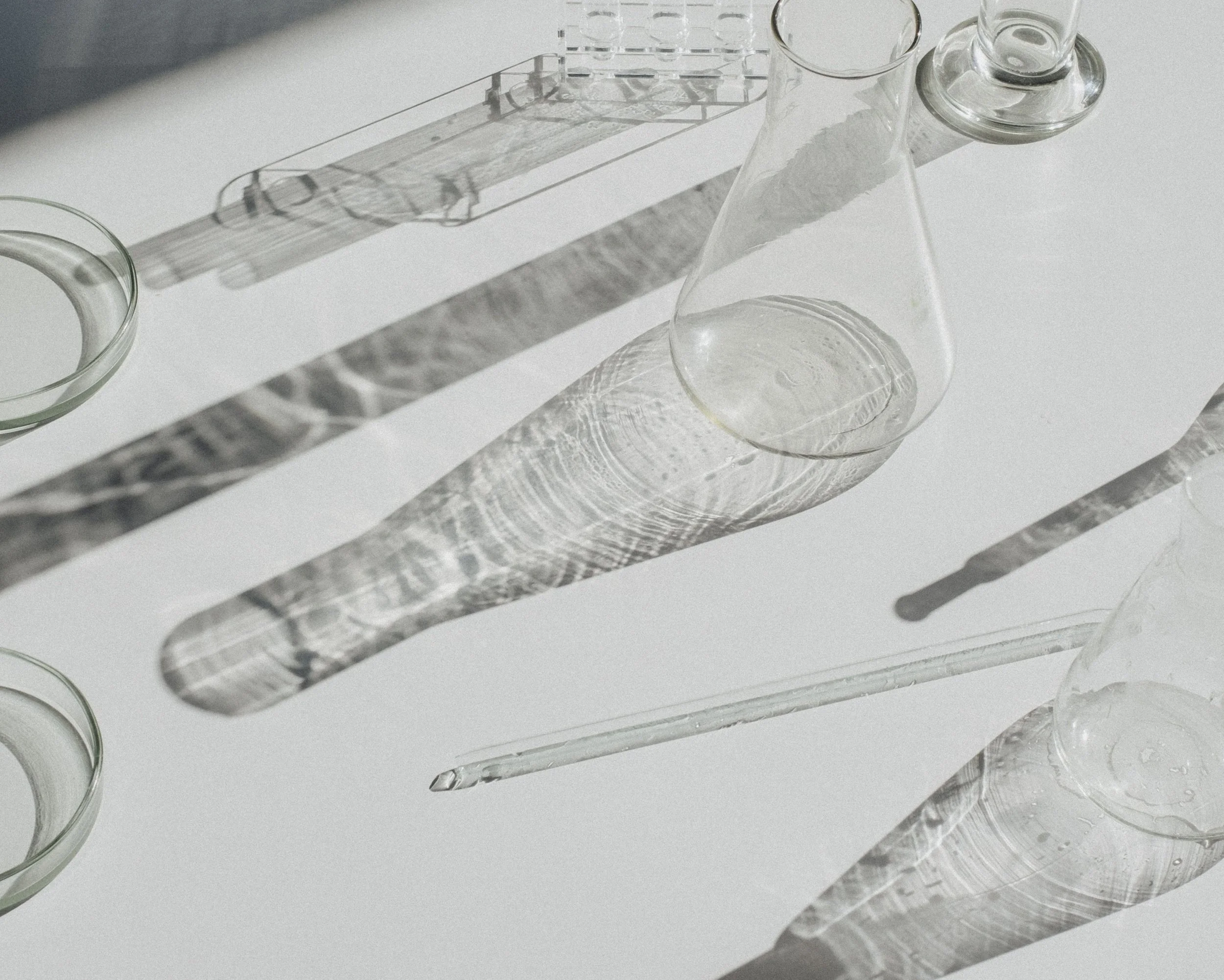 Glassware including a round bowl, a conical flask, and a beaker, casting shadows on a white surface with visible reflections and patterns.