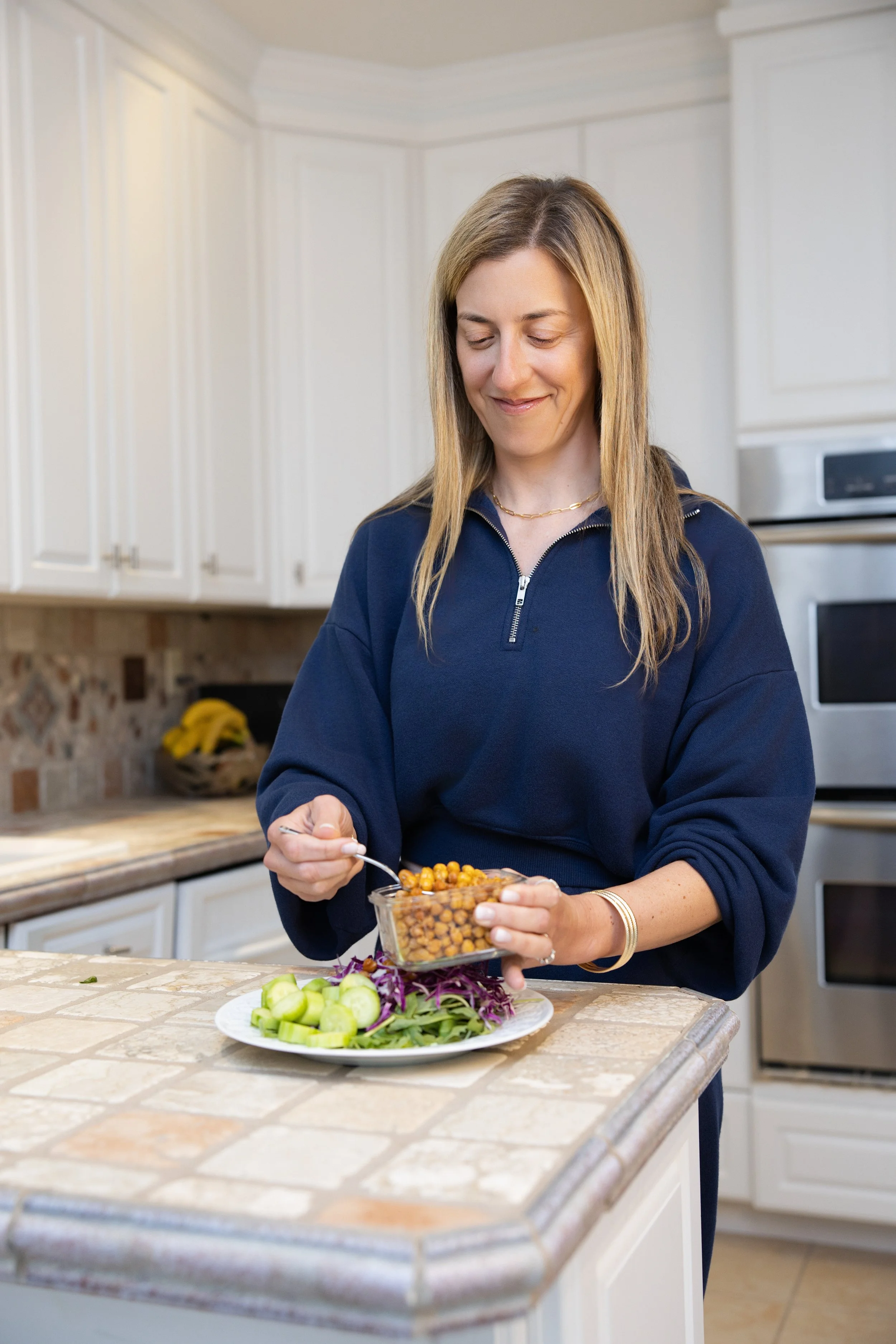 A woman with long blonde hair and a navy blue zip-up top, smiling softly while preparing a meal in a bright, white kitchen. She is holding a clear bowl of roasted chickpeas, standing in front of a marble countertop with a plate of sliced cucumbers, purple cabbage, and greens.