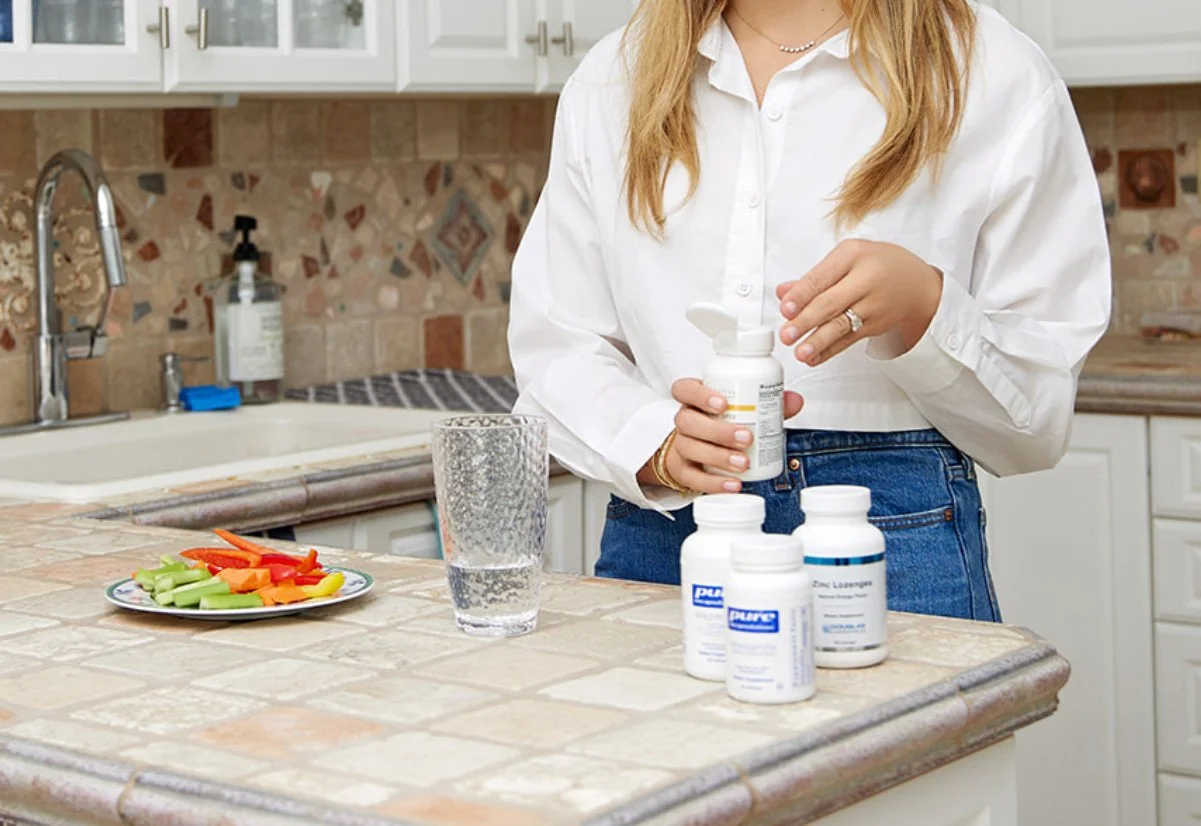 A woman in a white shirt and jeans standing in a kitchen, holding a pill bottle. On the counter are additional pill bottles and a glass of water, with a plate of sliced celery and carrots nearby.
