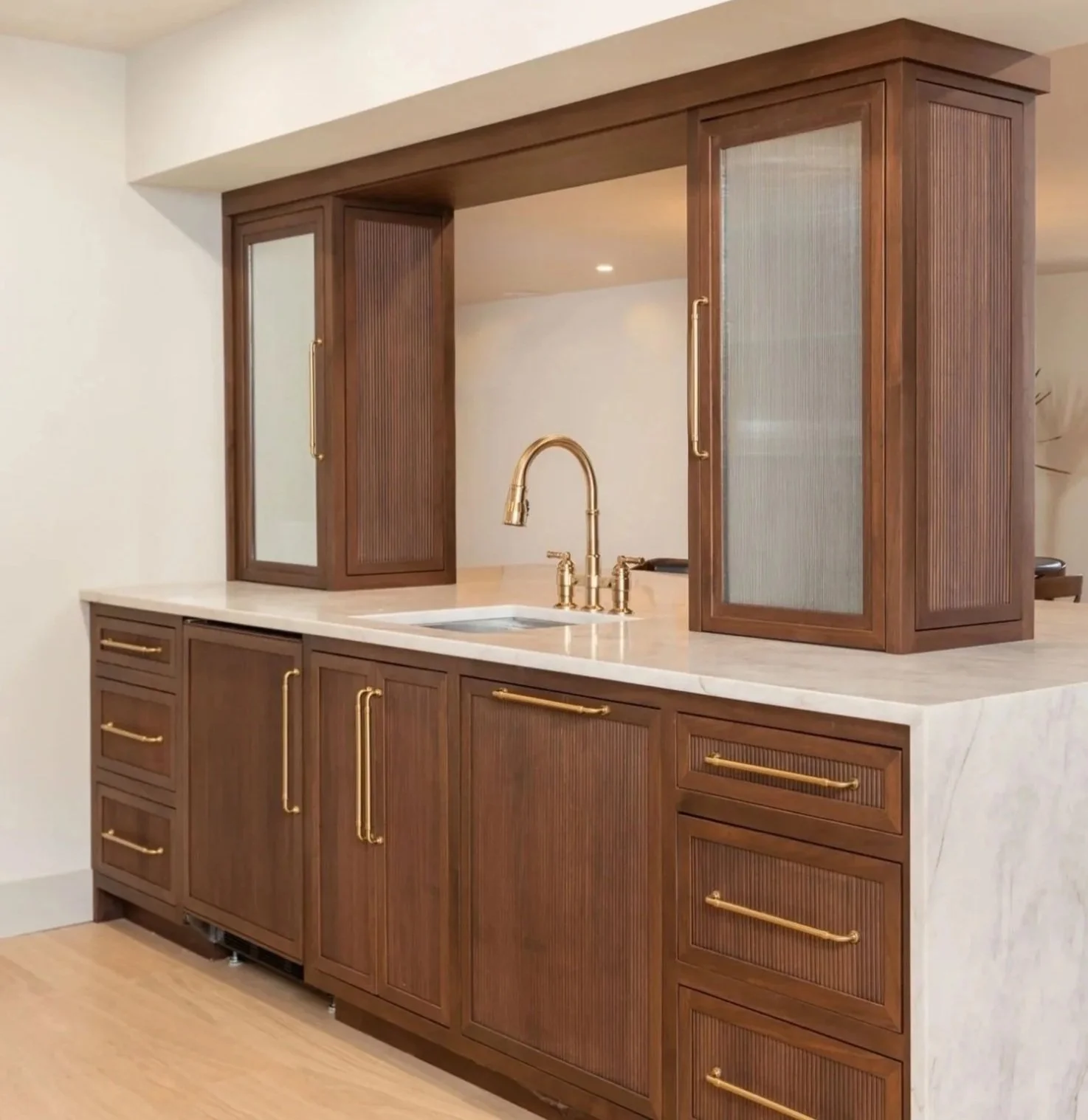 Fully custom walnut bar cabinetry with reeded door fronts, glass panels, brass hardware, and white stone countertop
