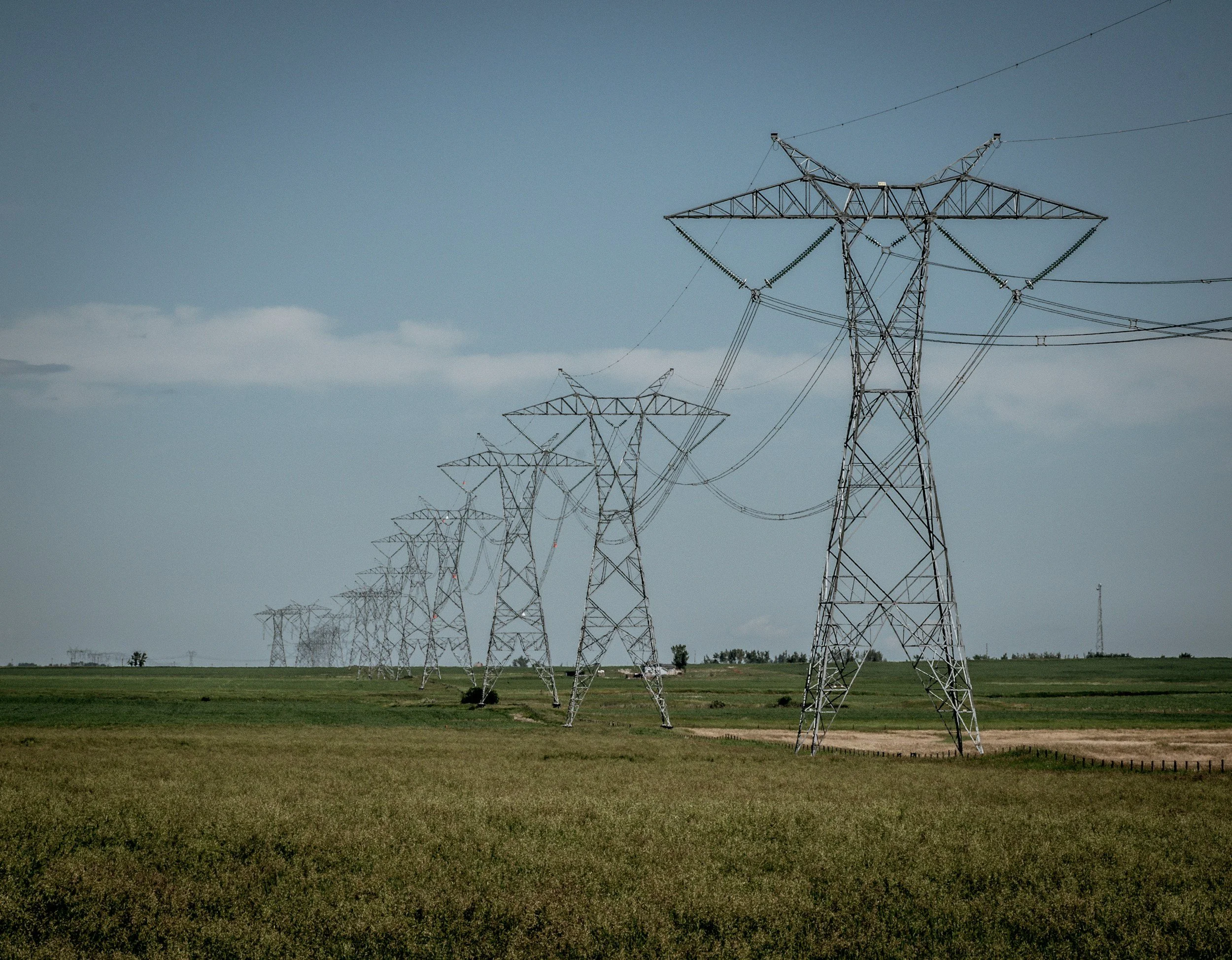 Multiple high-voltage power transmission towers across a grassy plain under a blue sky with some clouds.
