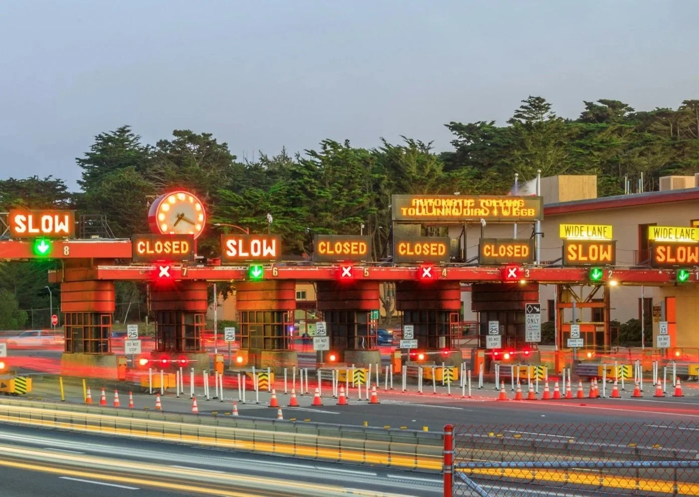 Toll booth with several lanes closed or slow, illuminated signals, and a digital sign with partial text, as traffic passes through.