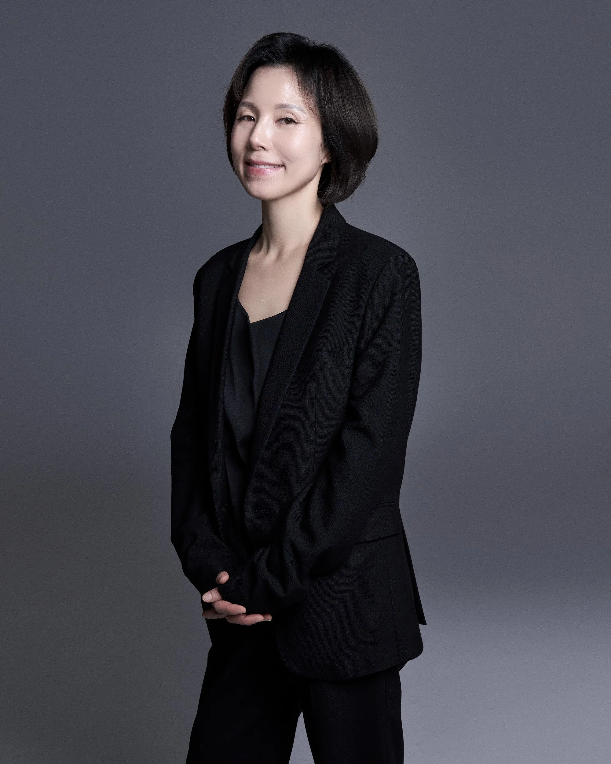 A professional woman with short dark hair wearing a black blazer and black blouse, standing against a gray background.