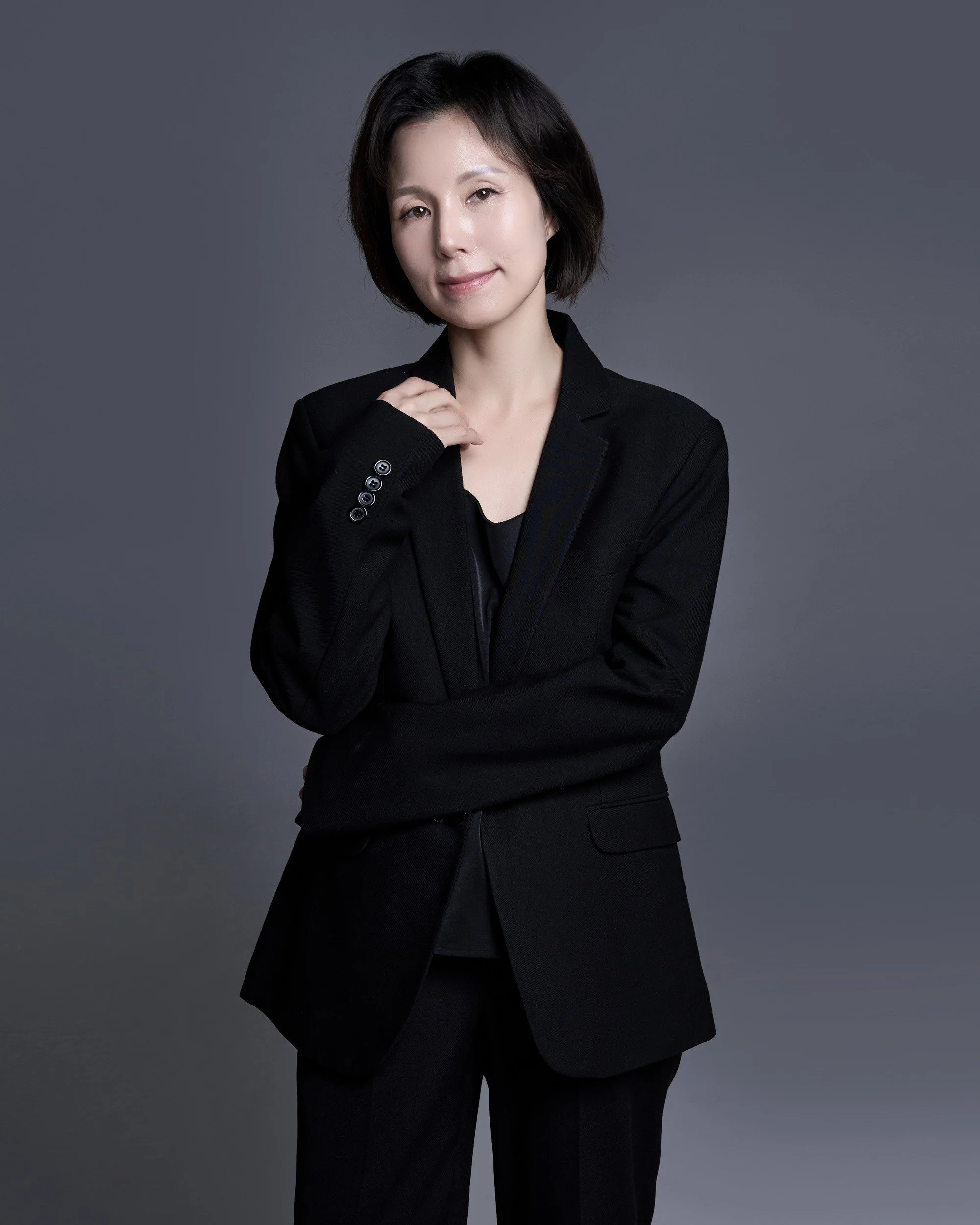 A woman wearing a black blazer and black pants posing against a dark gray background. She has short dark hair and a subtle smile.