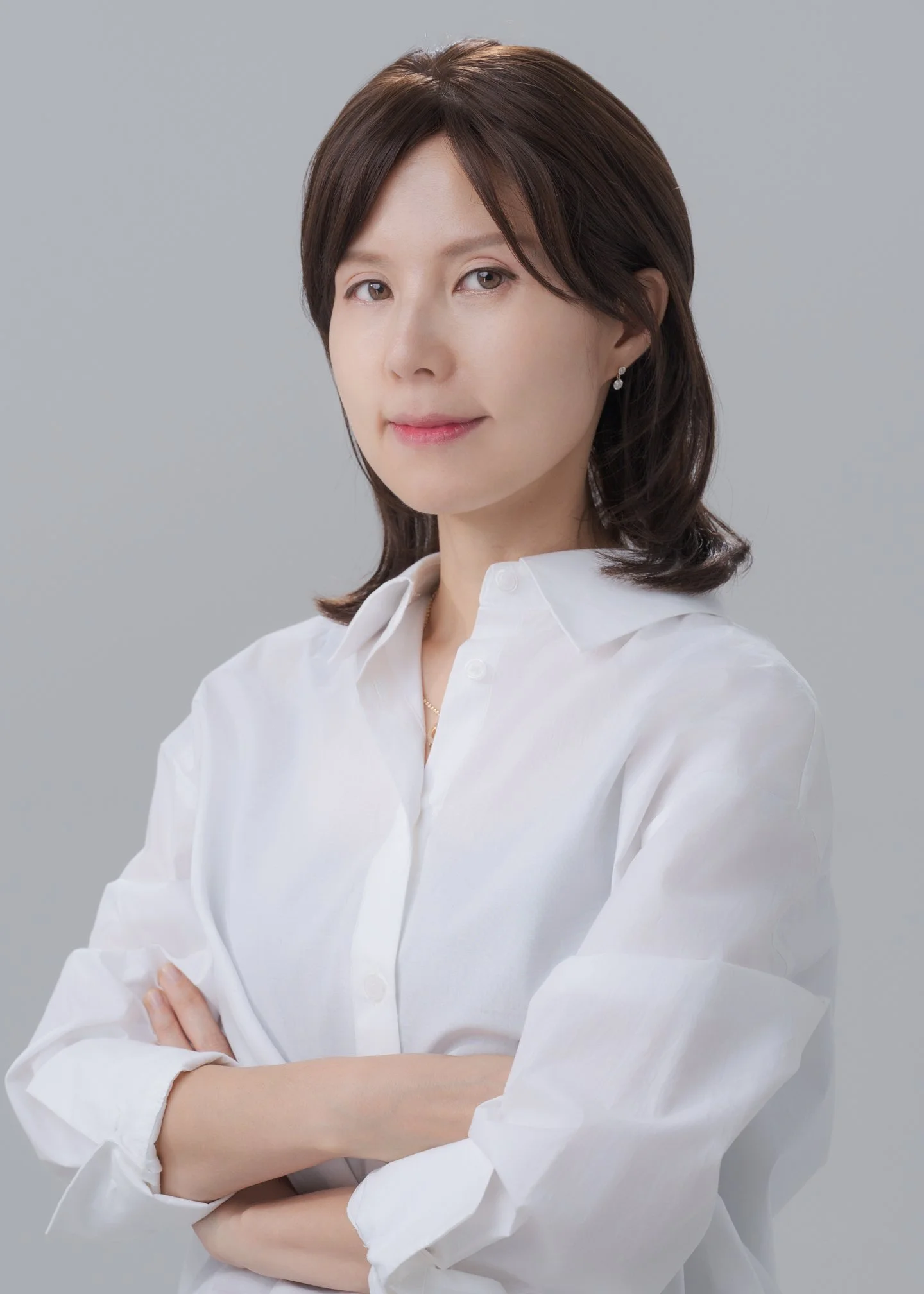 A woman with short brown hair wearing a white shirt and earrings, standing against a plain gray background.
