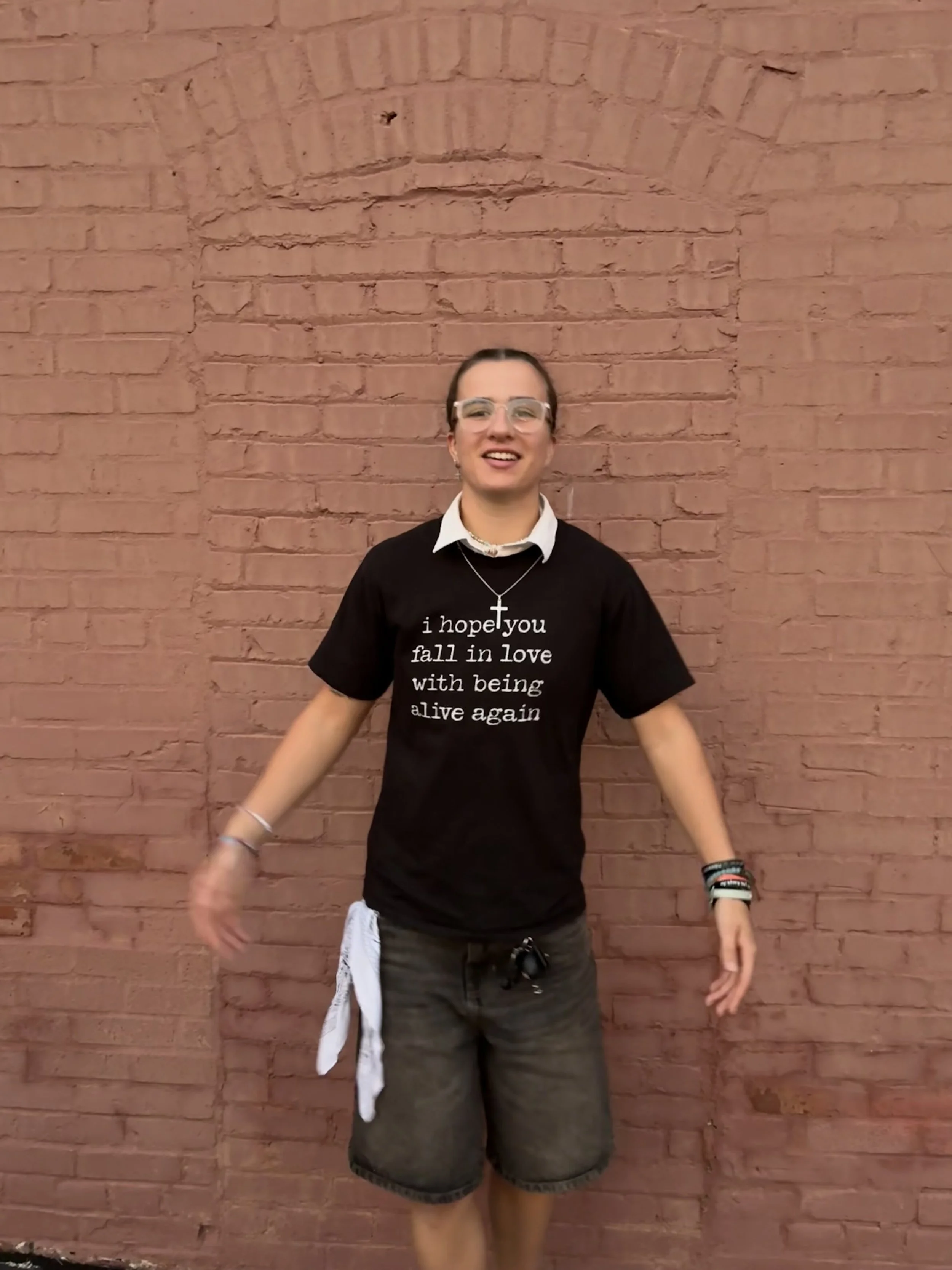 A person standing against a red brick wall, smiling, wearing glasses, a black t-shirt with white text, gray shorts, and accessories including bracelets, a necklace with a cross, and a white cloth hanging from the shorts.
