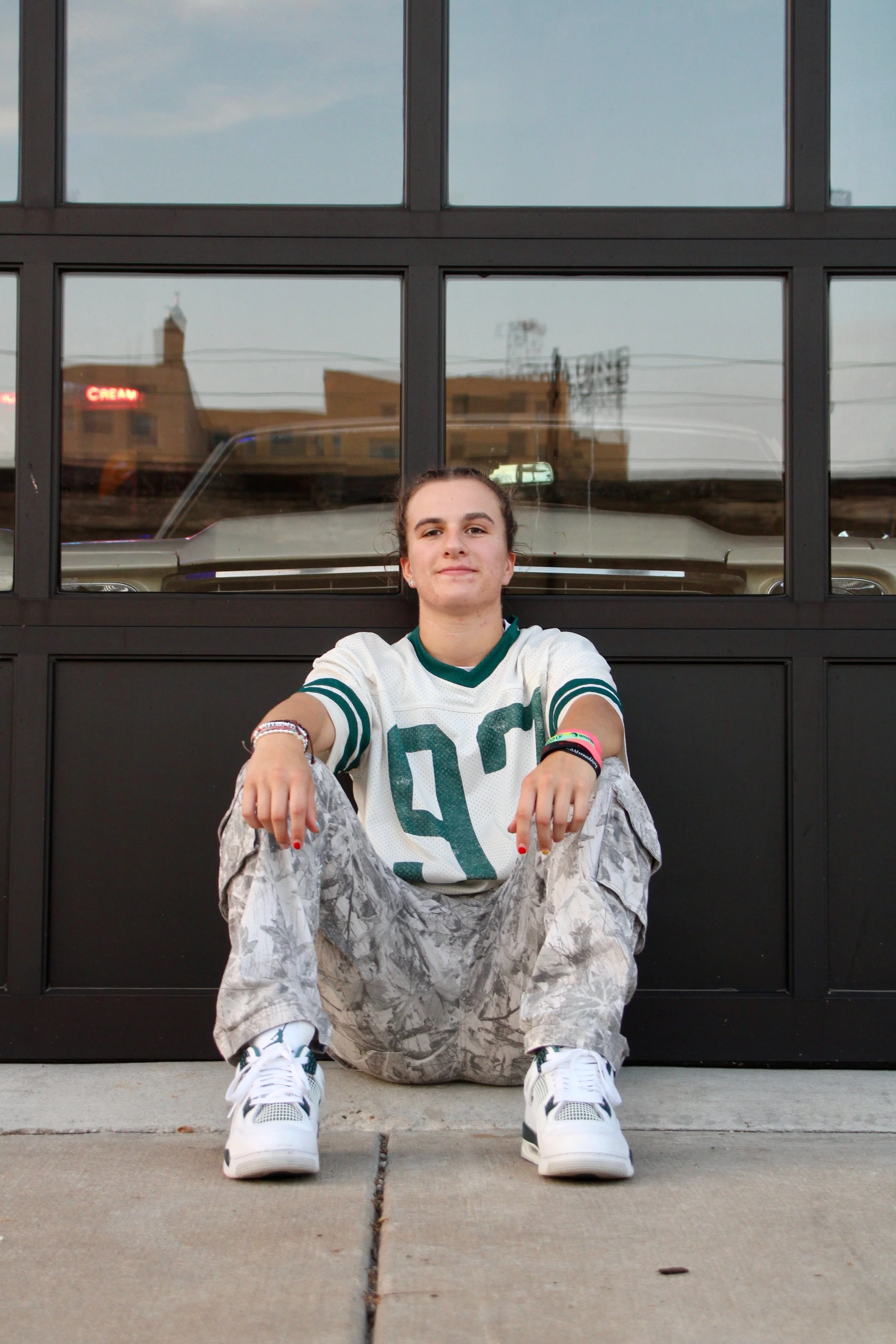 A young woman with a braids hairstyle sitting on the sidewalk in front of a black garage door, wearing a white sports jersey, camouflage pants, and white sneakers, with her arms resting on her knees, reflected in the glass windows of the garage door.